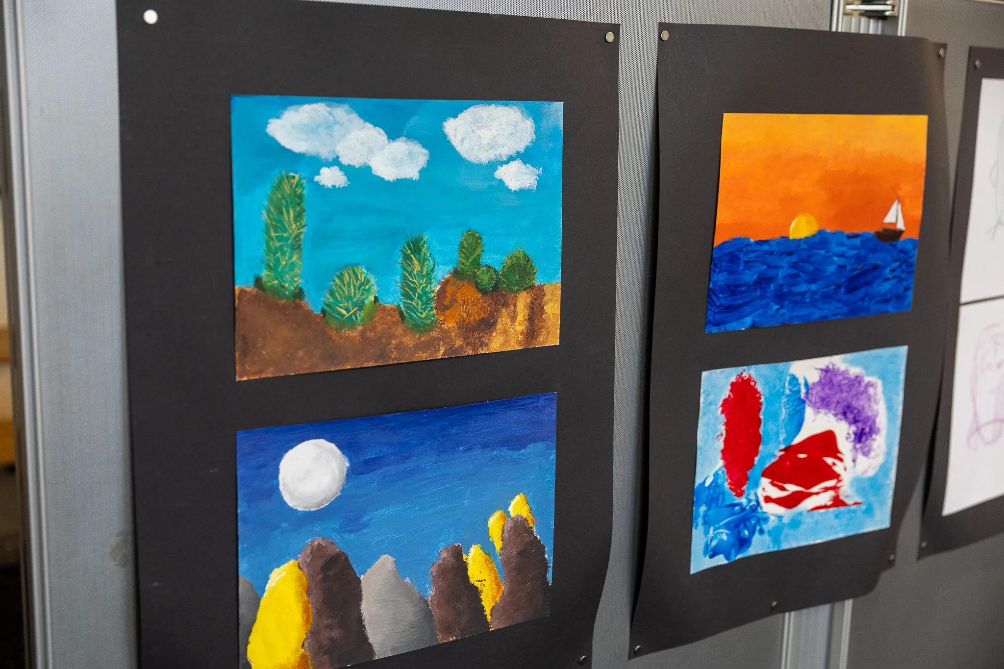 Vier Kindergemälde an einer schwarzen Wand hängen: eine Landschaft mit Bäumen und Wolken, ein Sonnenuntergang über dem Meer mit einem Segelboot, eine Berglandschaft mit Mond und ein abstraktes, farbenfrohes Bild.