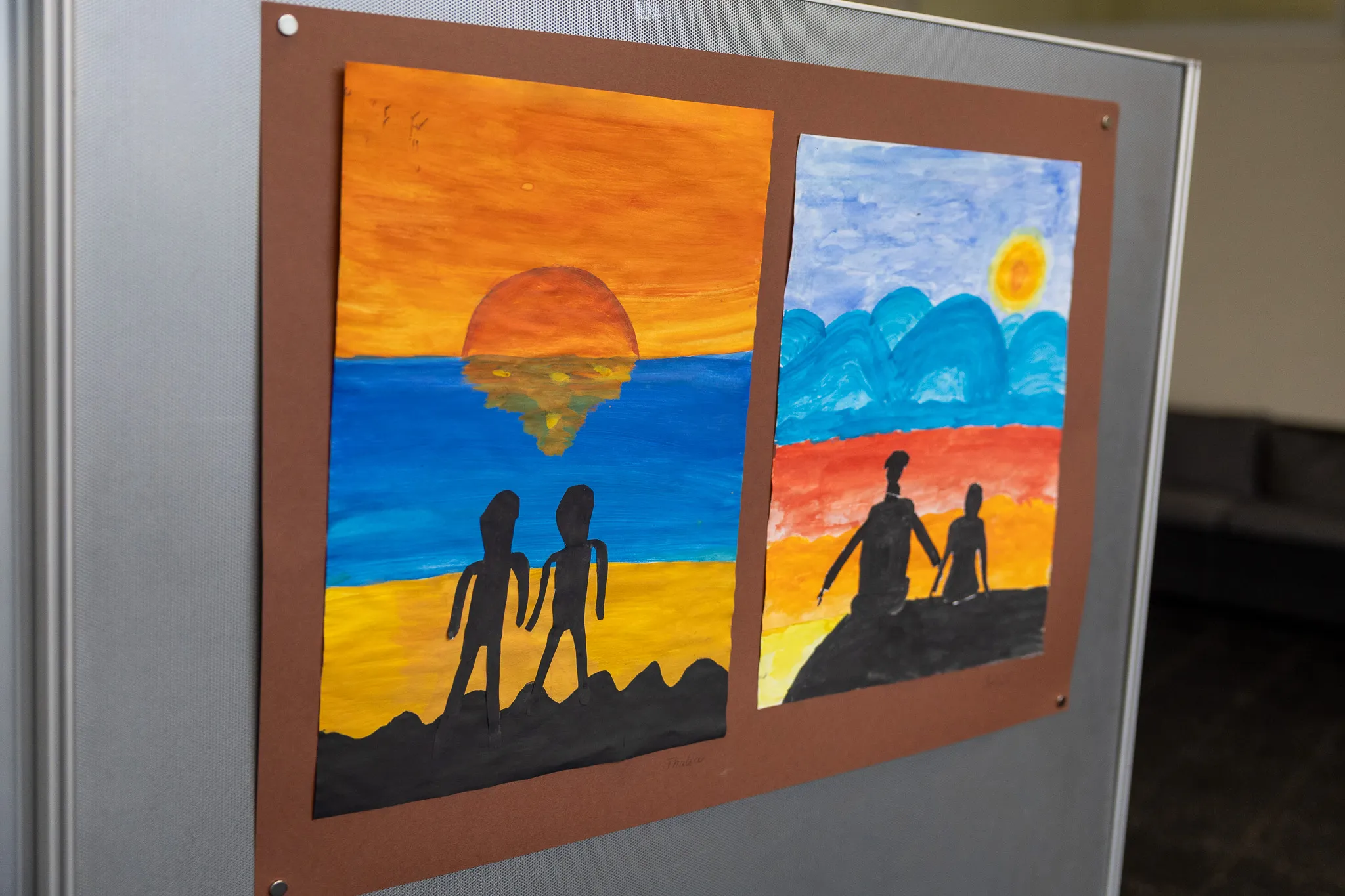 Zwei Kinderzeichnungen mit Silhouetten von Menschen vor Sonnenuntergängen hängen nebeneinander an einer grauen Wand.