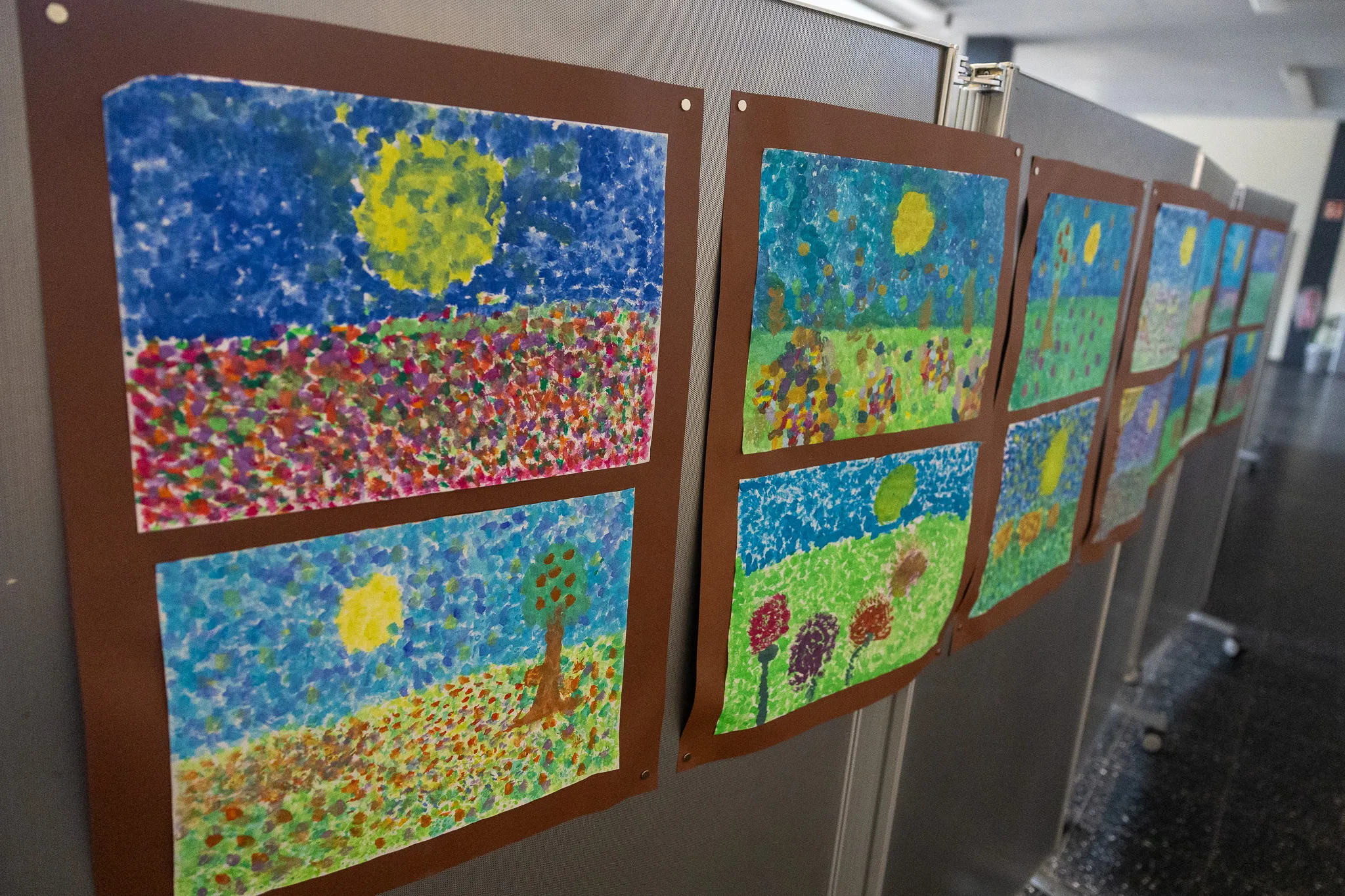 Mehrere Kinderzeichnungen mit Landschaftsmotiven hängen an einer grauen Wand in einer Ausstellung.