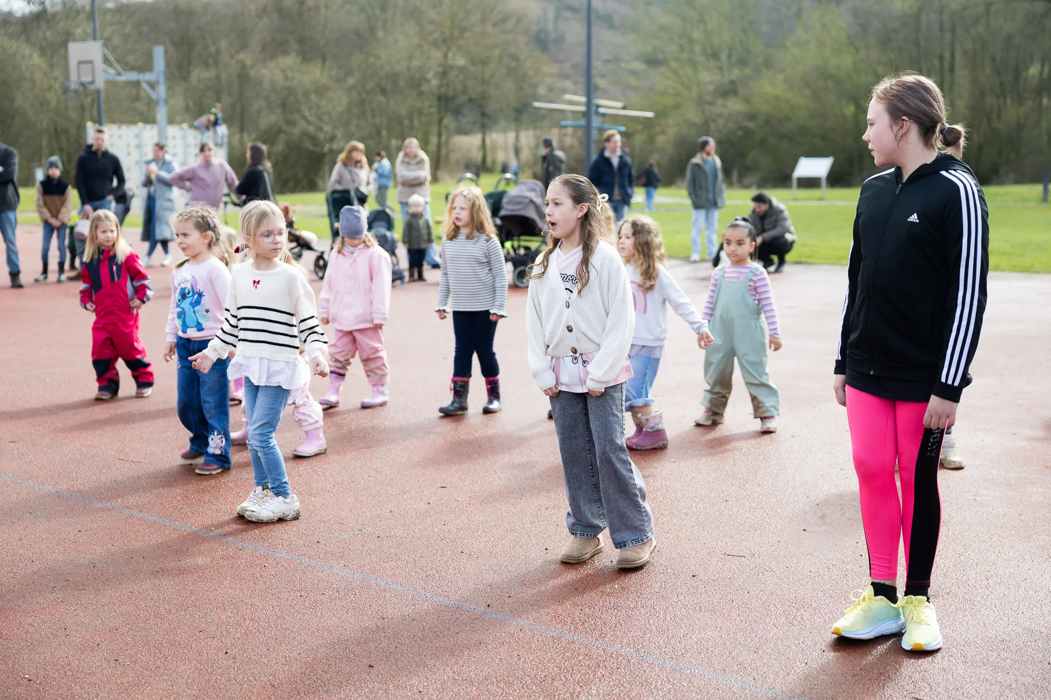 Eine Gruppe Kinder sucht auf einem roten Sportplatz nach Ostereiern, während ein Mädchen im Vordergrund Anweisungen gibt. Im Hintergrund stehen Eltern und beobachten.