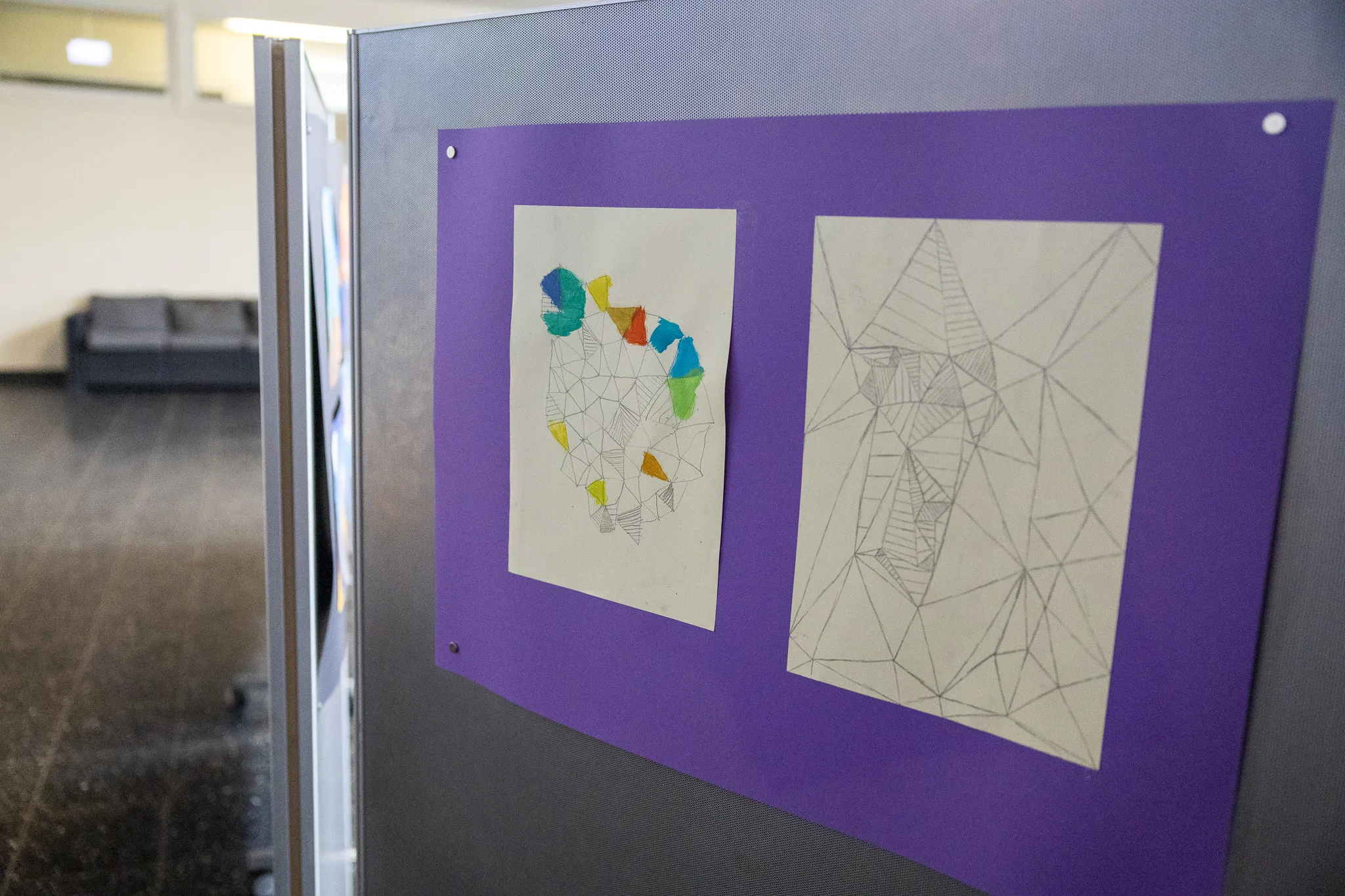 Zwei Zeichnungen auf Papier sind an einer grauen Wand auf einer violetten Platte befestigt. Die linke Zeichnung zeigt ein abstraktes Gesicht aus farbigen geometrischen Formen, die rechte eine abstrakte Komposition aus schwarzen Linien.