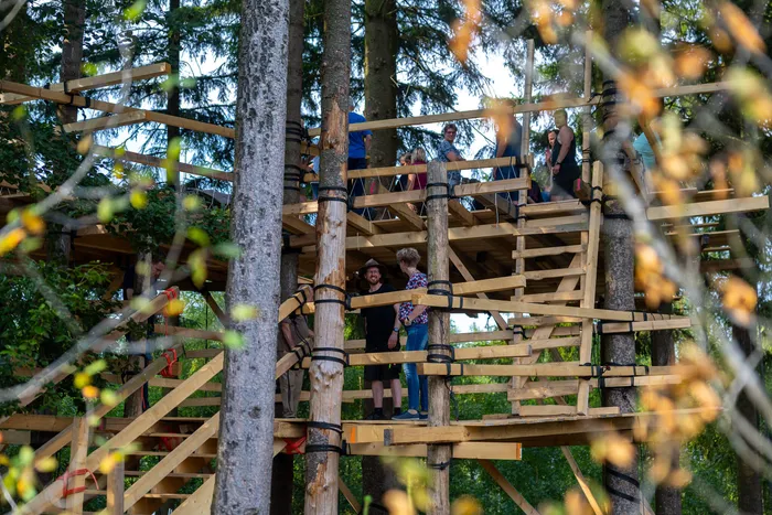 Mehrere Personen stehen auf den hölzernen Plattformen eines im Wald zwischen Bäumen errichteten Baumhauses.