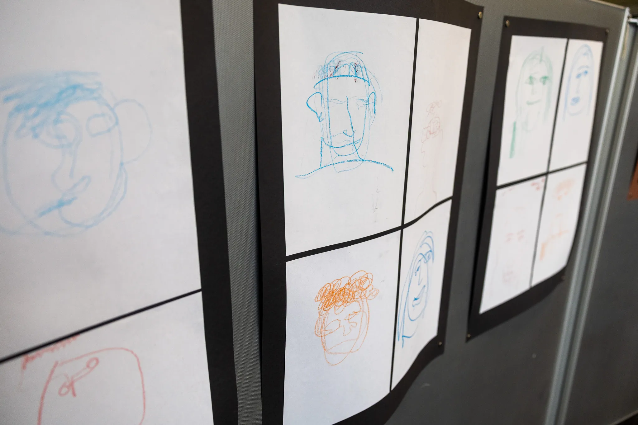 Mehrere Kinderzeichnungen von Gesichtern hängen in einer Galerie an einer grauen Wand.