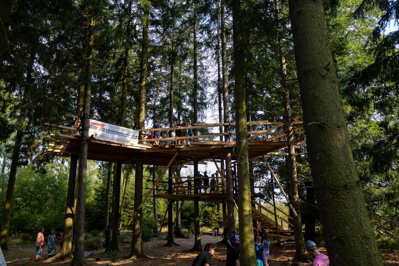 Ein mehrstöckiges Holzbaumhaus in einem hohen Nadelwald, auf dessen Plattformen Menschen stehen.