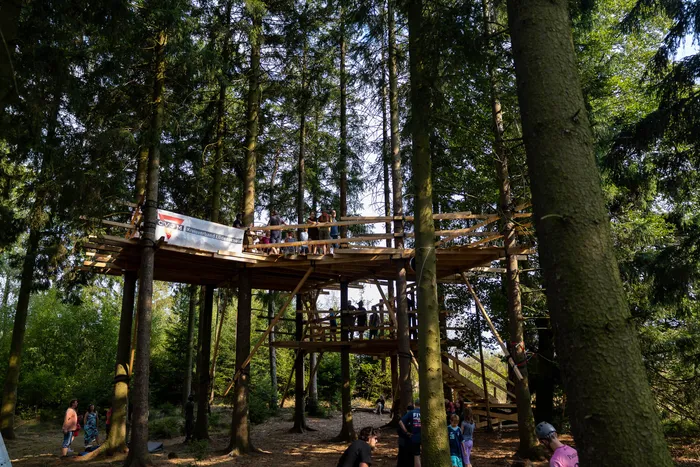 Ein mehrstöckiges Holzbaumhaus in einem hohen Nadelwald, auf dessen Plattformen Menschen stehen.