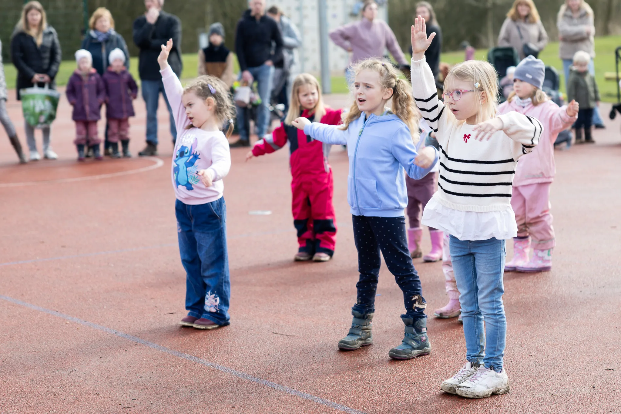 Mehrere Kinder mit erhobenen Händen stehen auf einem roten Sportplatz und nehmen an einer Ostereiersuche teil, im Hintergrund stehen weitere Personen.