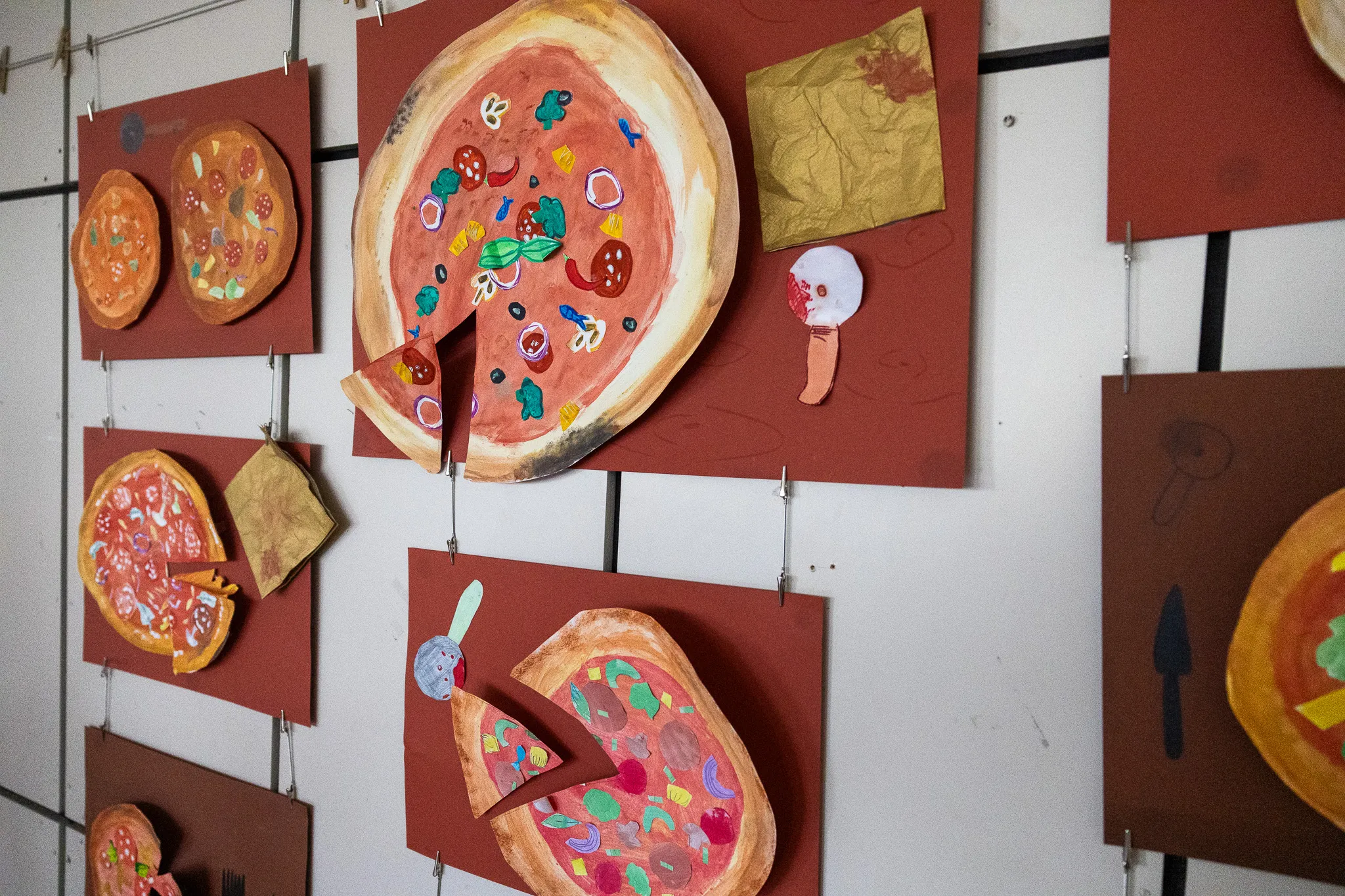 Mehrere bunte Pizzabilder, gefertigt von Kindern, hängen an einer weißen Wand in einer Ausstellung.