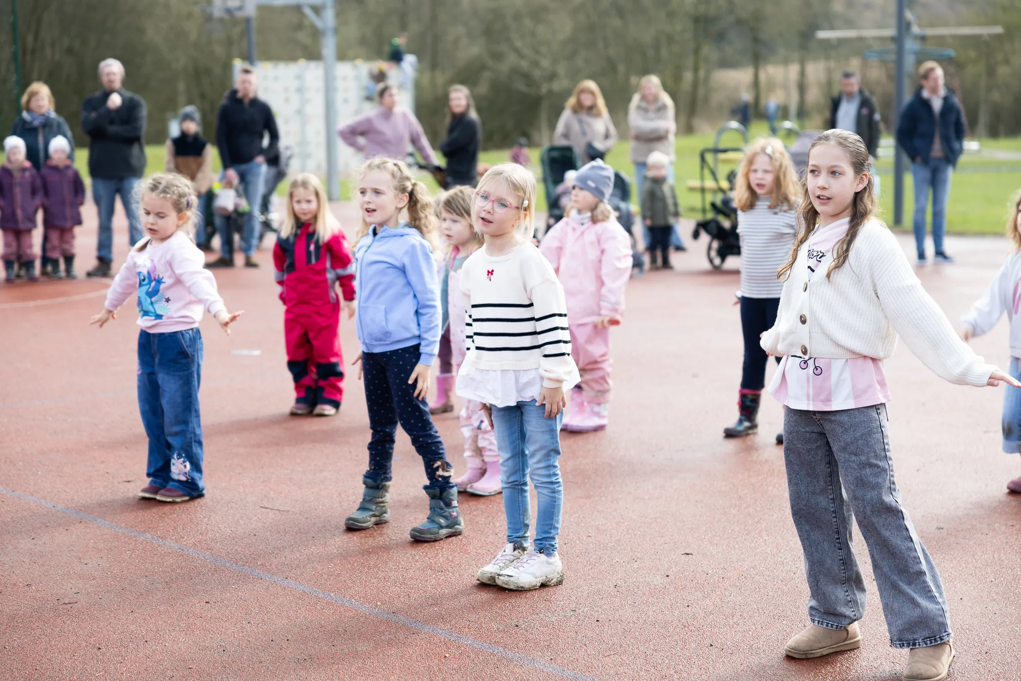 Eine Gruppe Kinder steht auf einem roten Sportplatz und streckt die Arme aus, während sie an einer Ostereiersuche teilnehmen. Im Hintergrund stehen Eltern und andere Zuschauer.