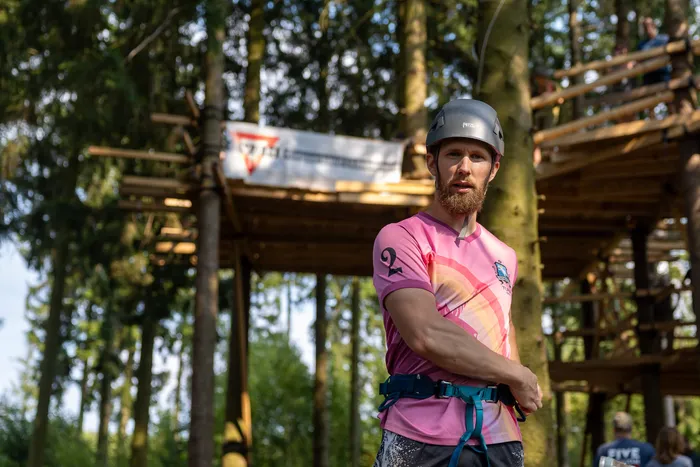 Ein bärtiger Mann mit Kletterhelm und Klettergurt steht vor einer hölzernen Konstruktion im Wald.