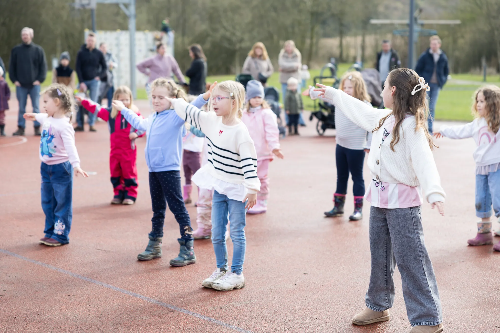 Kinder mit ausgestreckten Armen stehen auf einem roten Sportplatz und suchen nach Ostereiern. Im Hintergrund stehen weitere Familienmitglieder und Zuschauer.