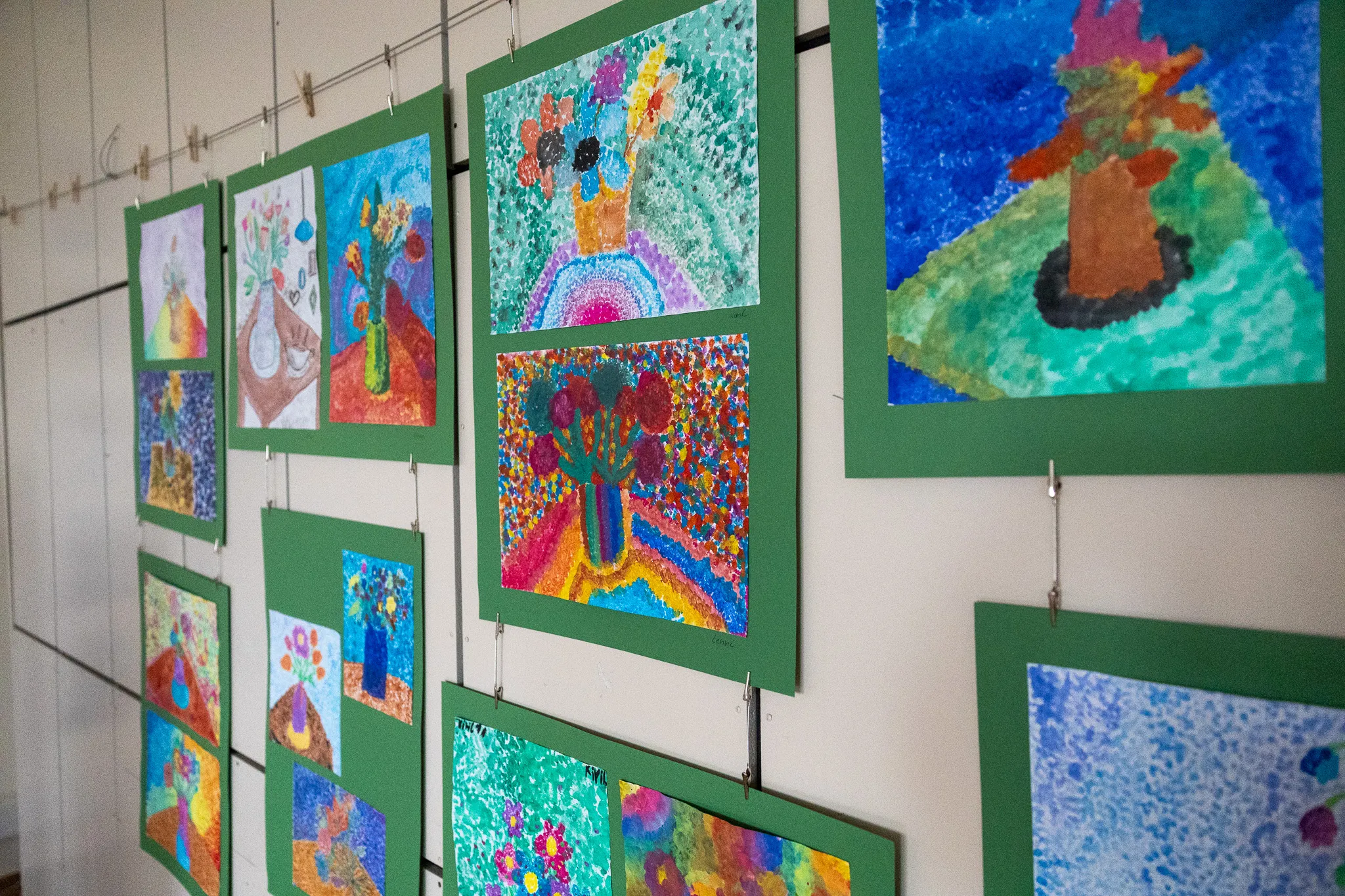 Mehrere farbenfrohe Kinderzeichnungen hängen an einer weißen Wand, präsentiert in grünen Rahmen und befestigt mit Wäscheklammern an einer Schnur.