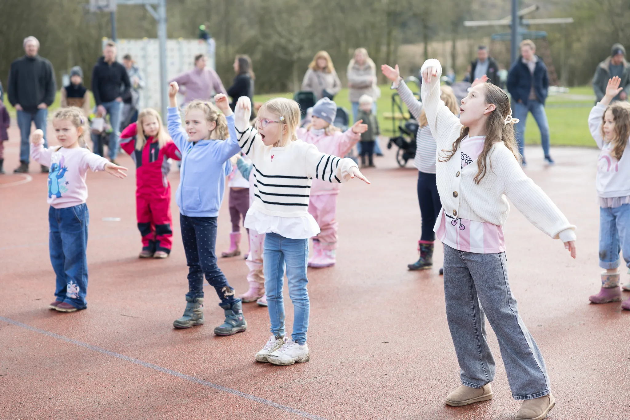 Eine Gruppe Kinder mit erhobenen Armen tanzt oder spielt auf einem roten Sportplatz, während im Hintergrund Eltern zusehen.