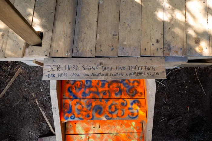 Ein handbeschriebenes Holzschild mit einem Segensspruch über bunt bemalten Holzbrettern eines Baumhauses.
