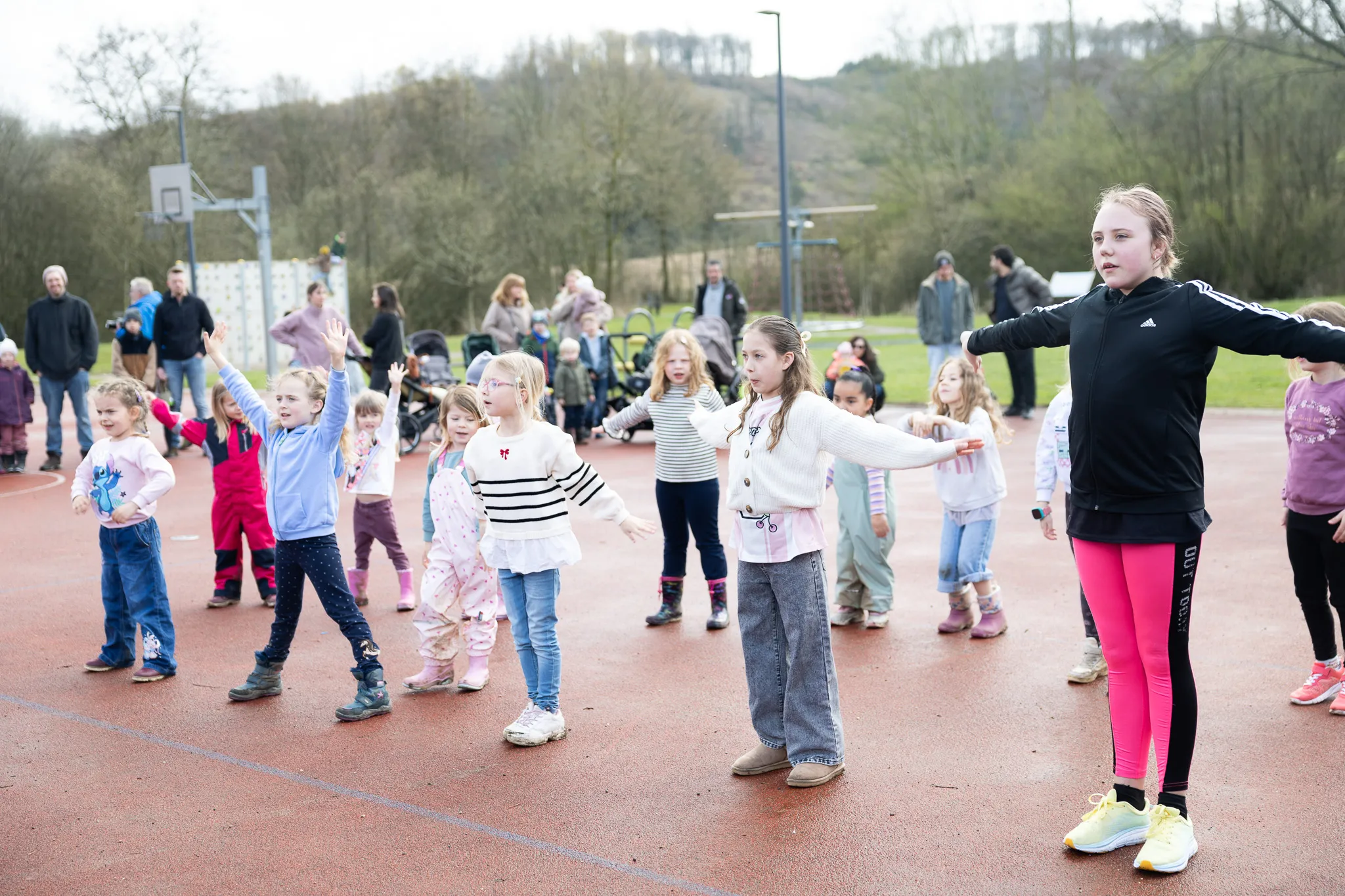 Eine Gruppe Kinder steht auf einem roten Sportplatz und streckt die Arme seitlich aus, während sie an einer Aktivität teilnehmen. Im Hintergrund stehen weitere Familienmitglieder und ein Basketballkorb.