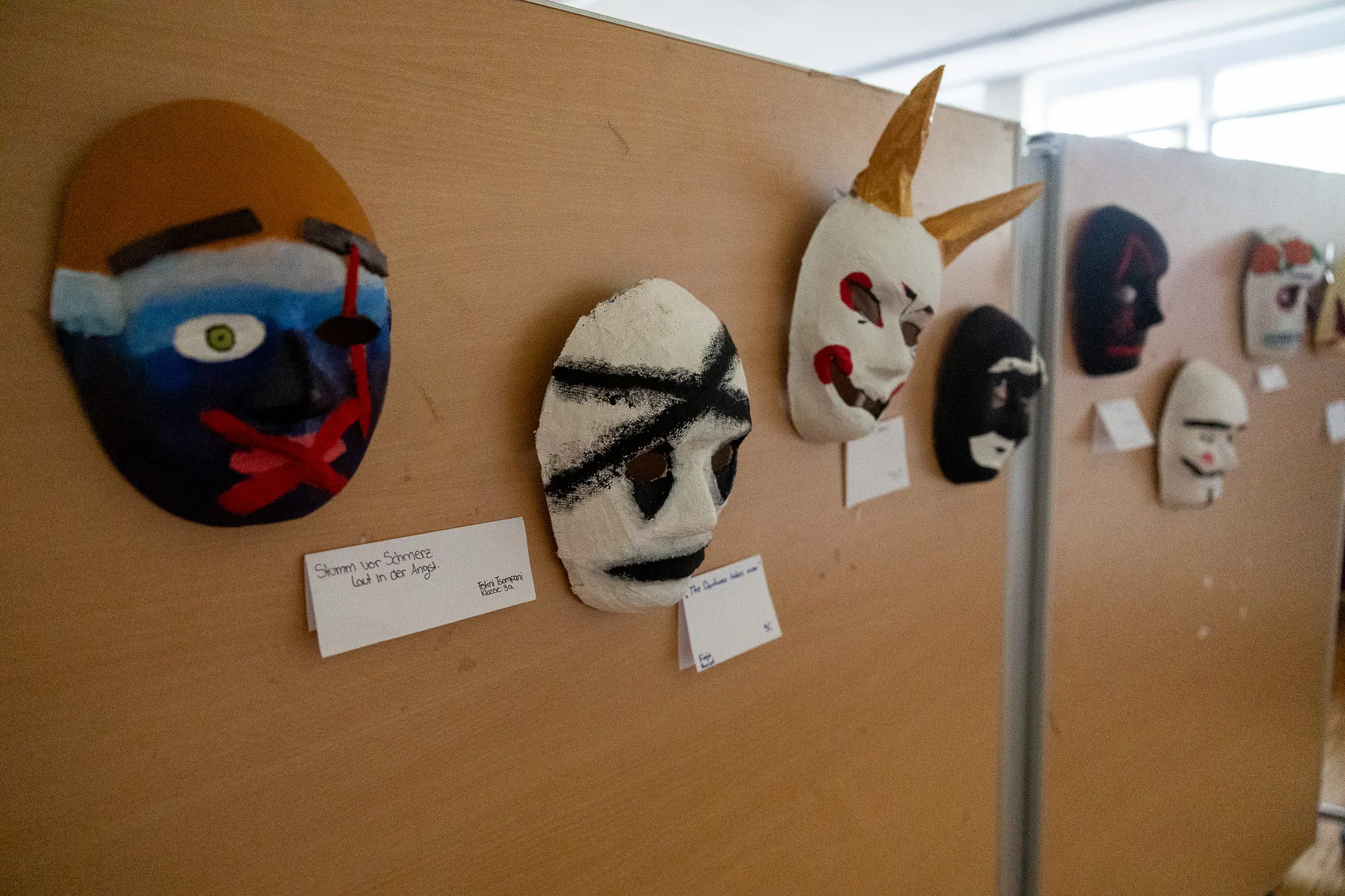 Mehrere bemalte Masken hängen an einer hellbraunen Wand in einer Ausstellung. Beschriftungen geben Informationen zu den Kunstwerken und ihren Schöpfern an.