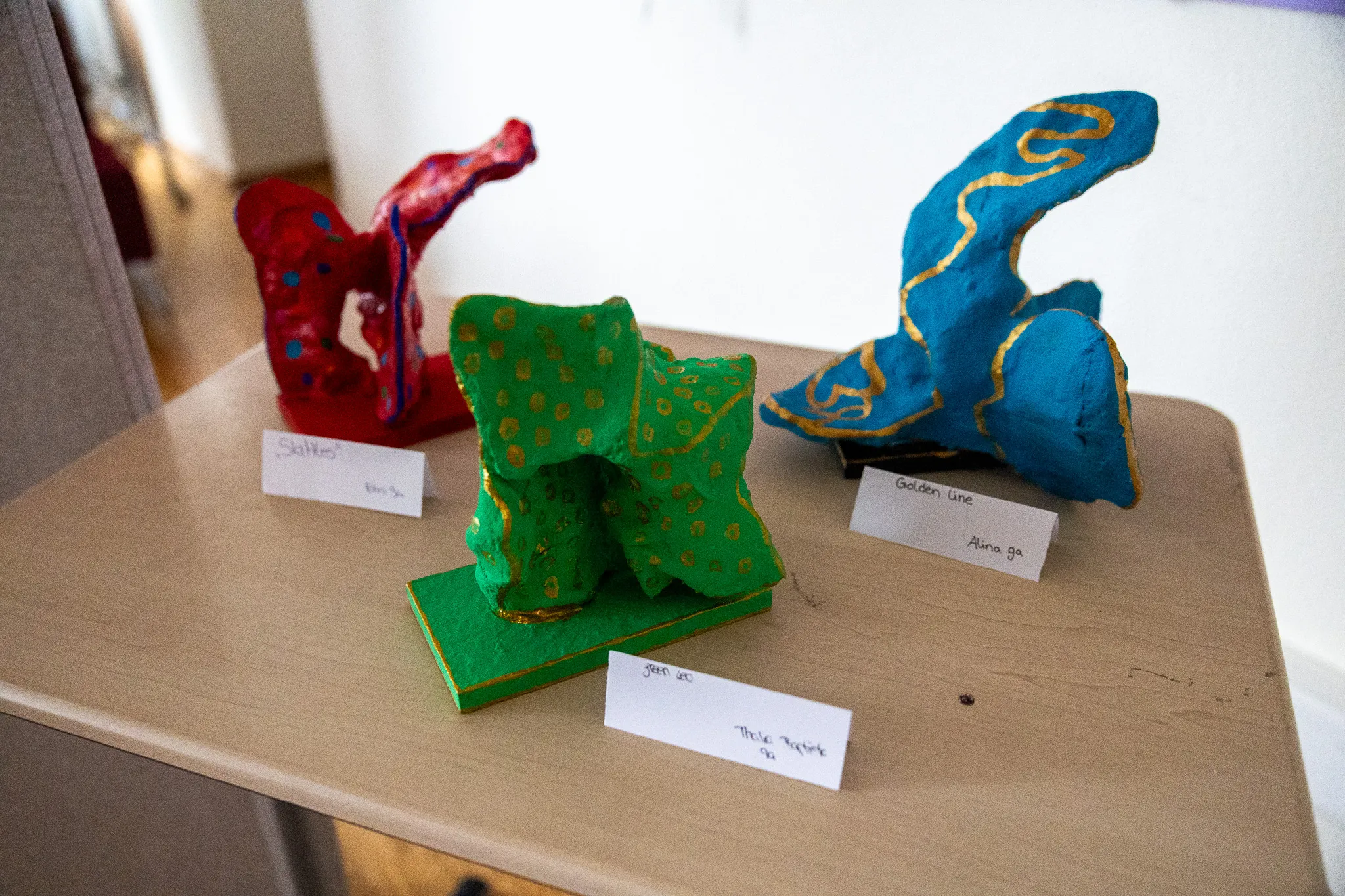 Drei farbige Tonfiguren stehen auf einem Tisch, beschriftet mit den Namen der Schüler, die sie gestaltet haben.