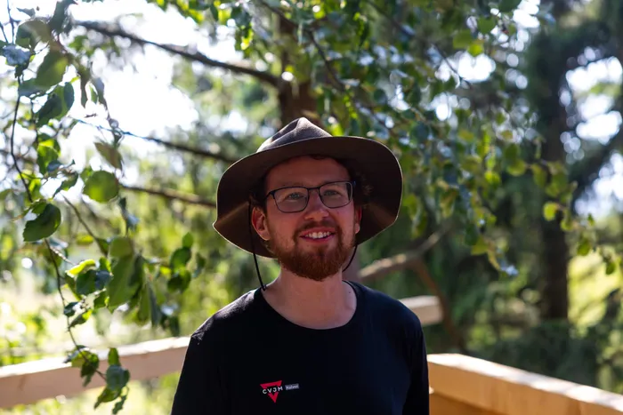 Ein junger Mann mit Brille und braunem Hut lächelt vor einem Hintergrund aus grünen Bäumen und einer Holzkonstruktion.