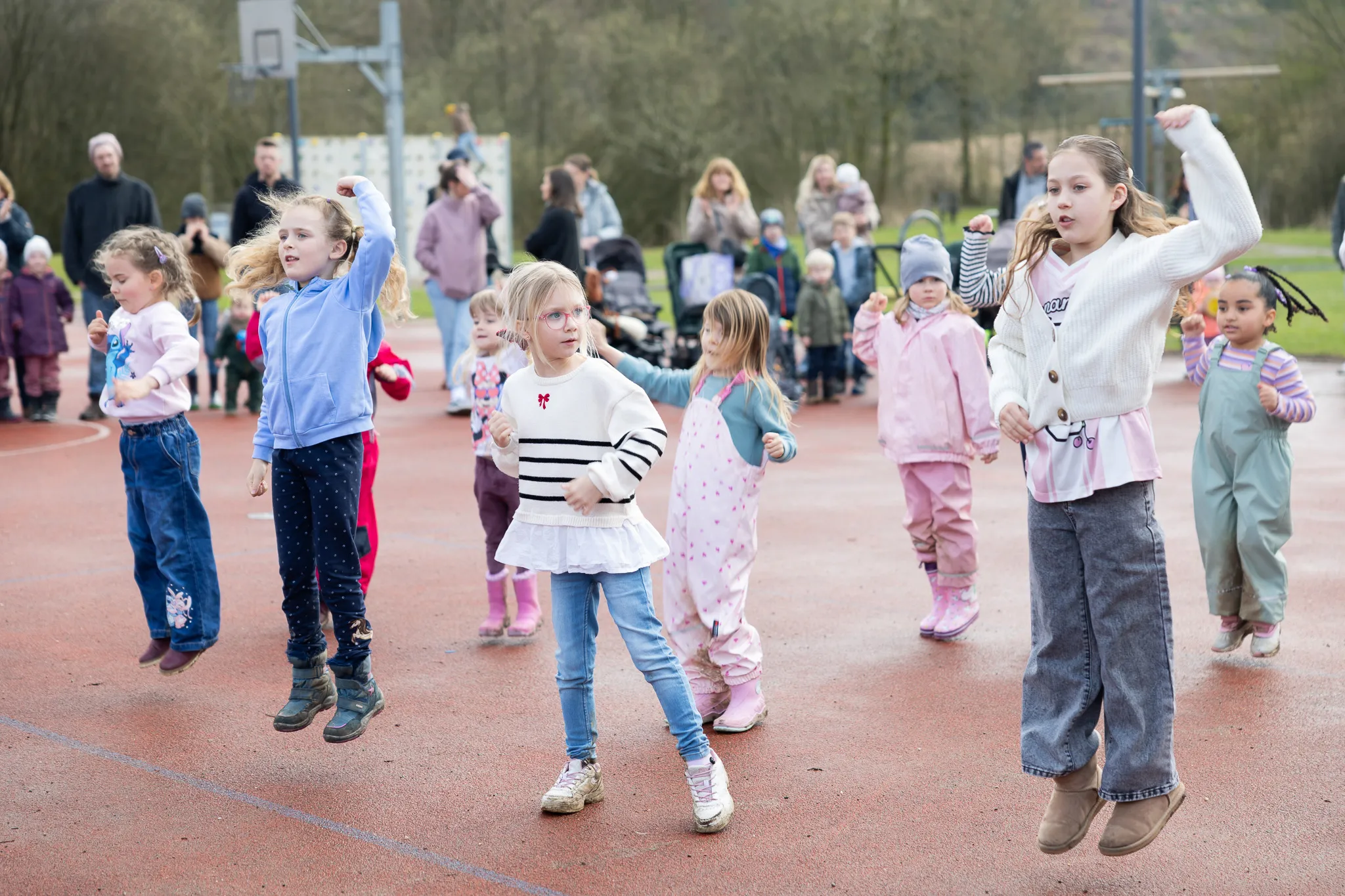Mehrere Kinder springen und laufen auf einem roten Sportplatz, während im Hintergrund weitere Familienmitglieder stehen. Im Fokus eine Gruppe Mädchen bei einer Ostereiersuche.
