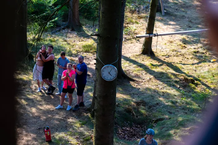 Eine Gruppe von Jugendlichen steht auf einem Waldweg im Wald; an einem Baum hängt eine Uhr.