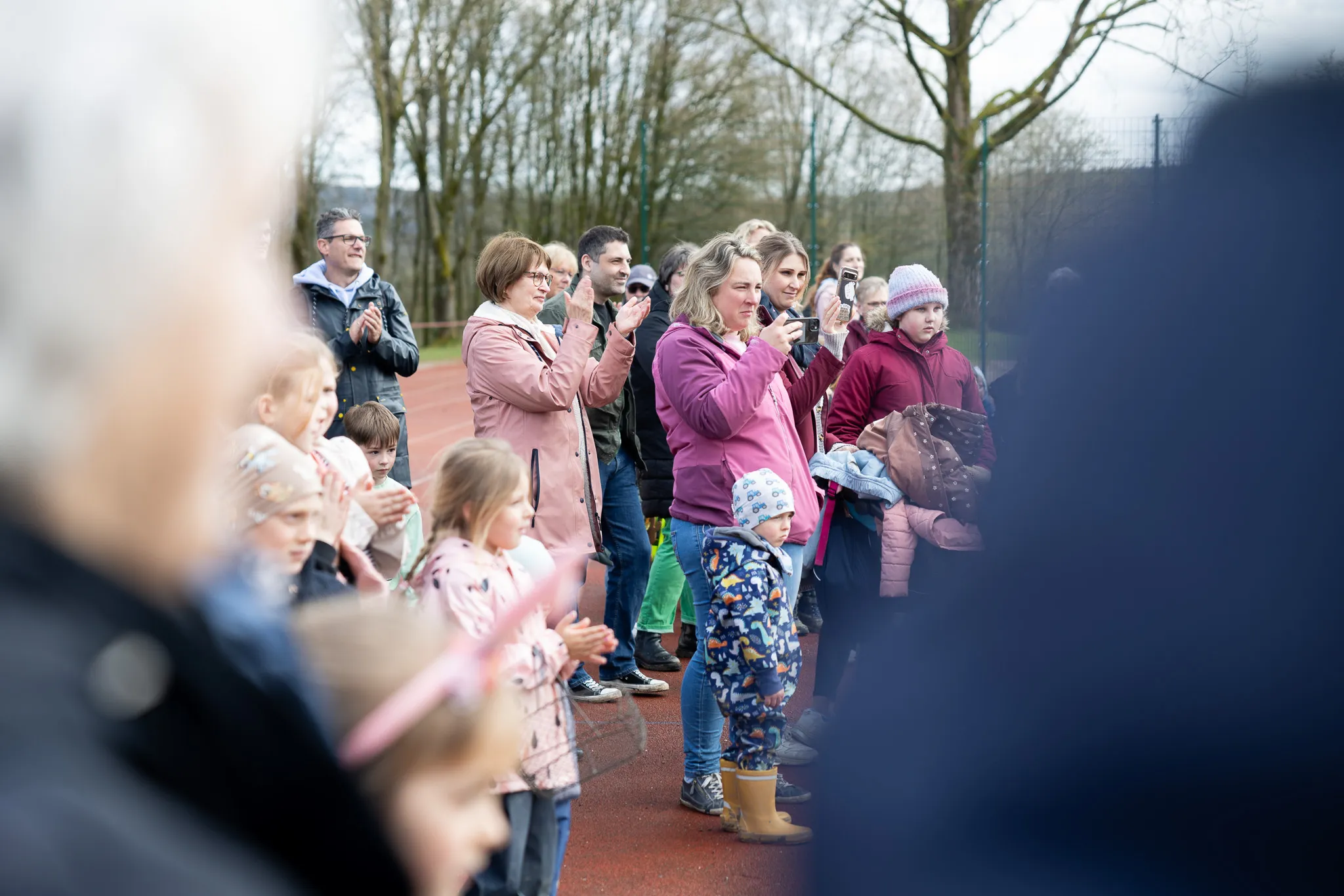 Zuschauer, darunter viele Kinder, stehen an einem roten Sportplatz und beobachten ein Ereignis. Einige halten Smartphones hoch, um Fotos oder Videos aufzunehmen.
