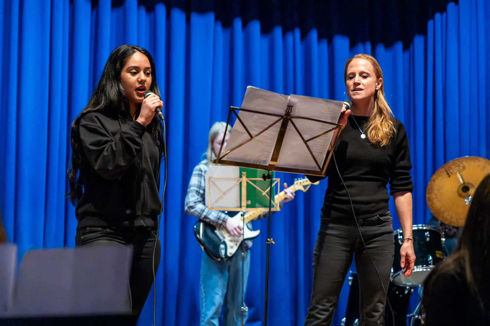 Zwei Frauen singen auf einer Bühne vor blauem Hintergrund in die Mikrofone, während im Hintergrund ein Gitarrist und Schlagzeuger spielen.