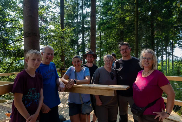 Eine Gruppe von Menschen steht lächelnd auf einer Holzkonstruktion in einem Wald.