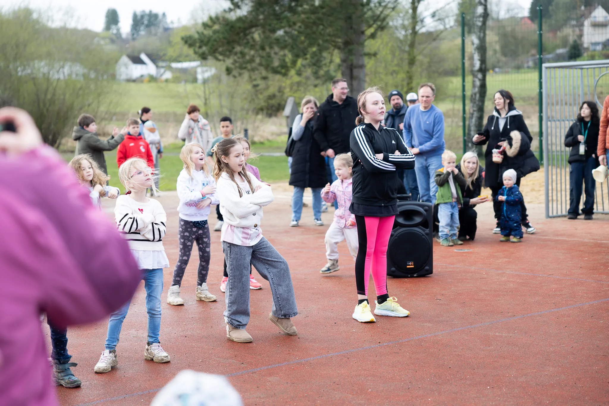Eine Gruppe Kinder tanzt und spielt auf einem roten Sportplatz, während Eltern und andere Zuschauer zusehen. Im Hintergrund sind Bäume und Häuser zu erkennen.