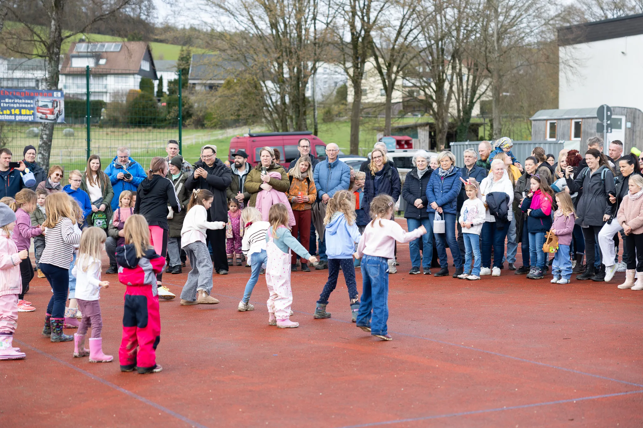 Kinder tanzen im Kreis, umgeben von einer großen Gruppe zuschauender Erwachsener auf einem roten Belag. Im Hintergrund sind Häuser und Bäume zu sehen.