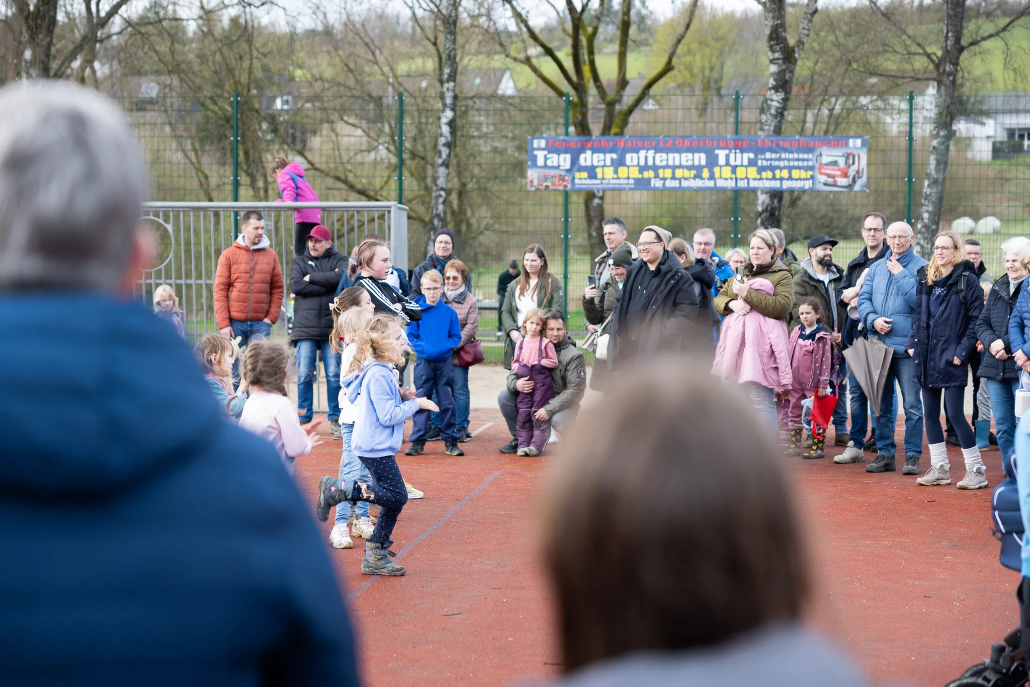 Kinder suchen auf einem roten Platz nach Ostereiern, umringt von zuschauenden Eltern und anderen Besuchern. Im Hintergrund ein Banner mit der Aufschrift „Tag der offenen Tür“.