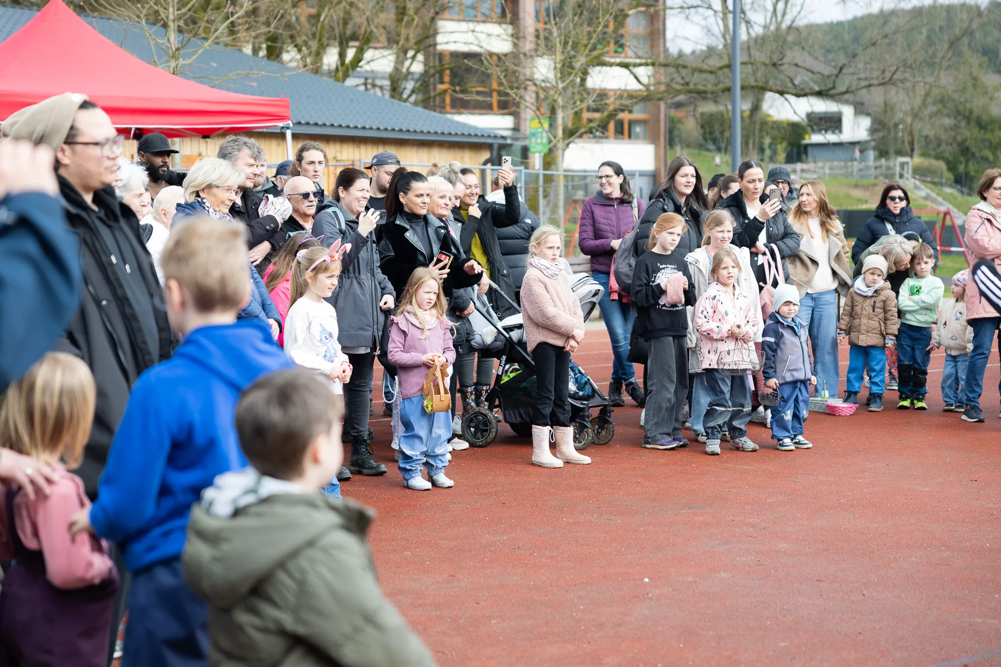 Zuschauer beobachten eine Ostereiersuche auf einem roten Sportplatz, viele halten Smartphones hoch.