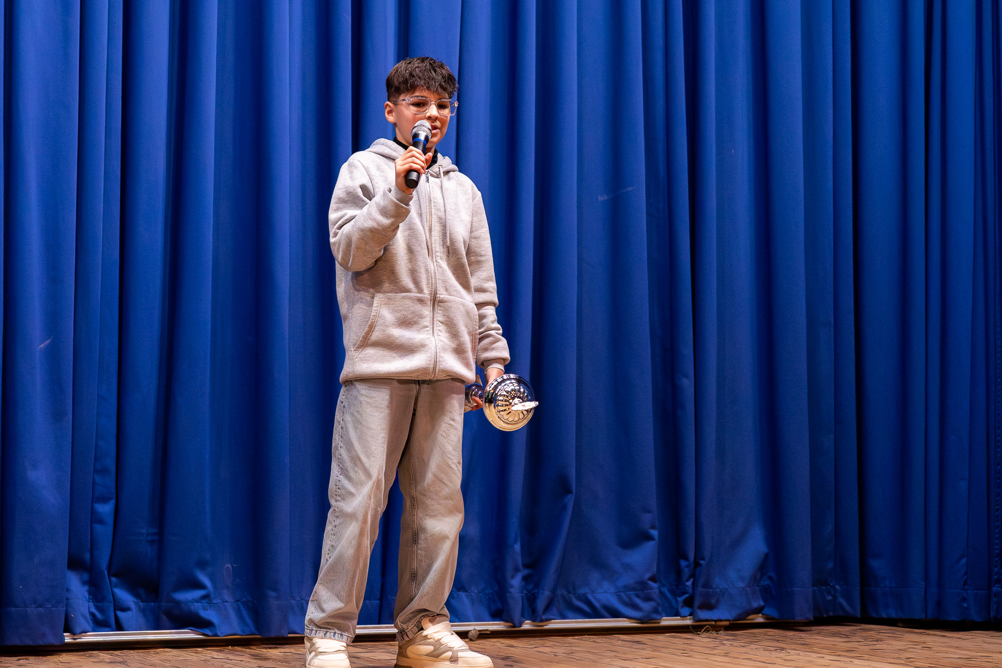 Ein Junge mit Brille hält ein Mikrofon und eine Auszeichnung in der Hand, während er auf einer Bühne vor einem blauen Vorhang spricht.