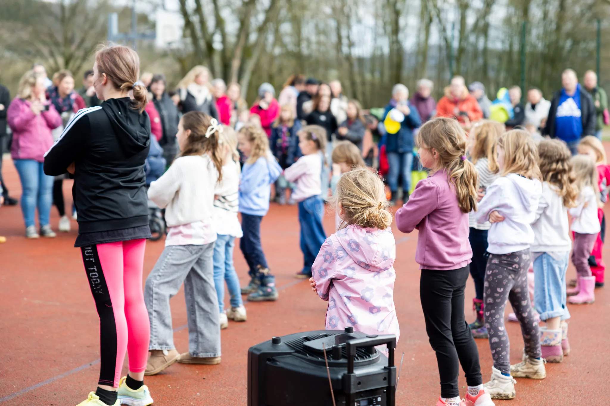 Kinder stehen im Kreis auf einem roten Sportplatz und halten Hände, während im Hintergrund Zuschauer stehen. Eine Lautsprecheranlage steht vor den Kindern.