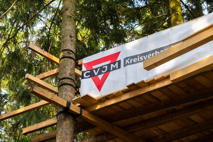 Ein Banner mit dem CVJM-Logo ist an der Holzkonstruktion eines Baumhauses in einem Wald angebracht.