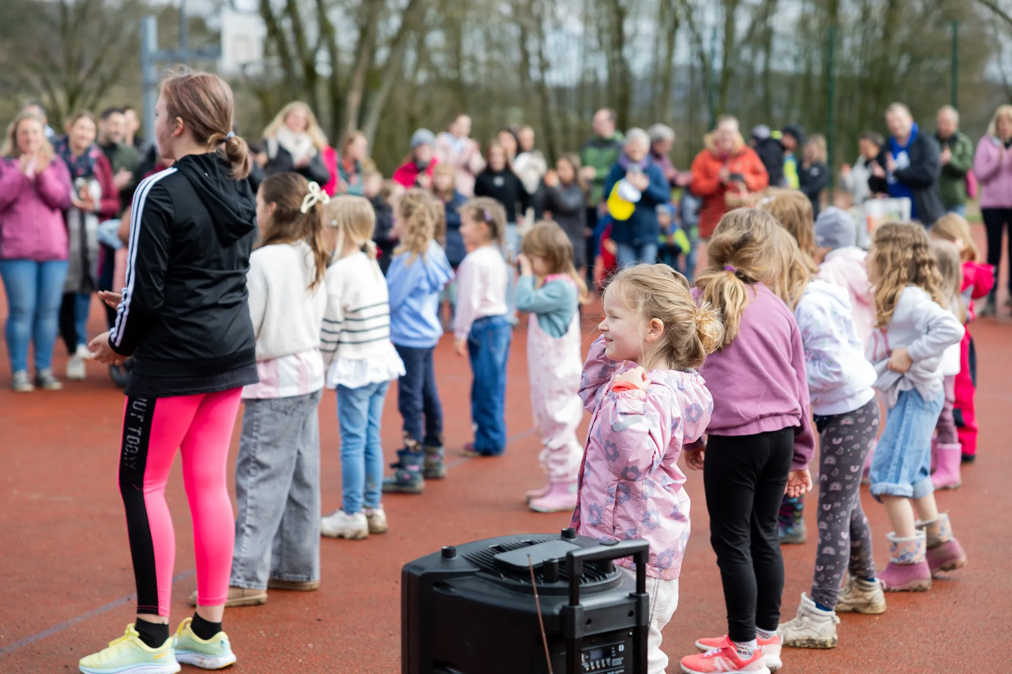 Eine Betreuerin steht vor einer Gruppe Kinder, die sich an den Händen halten und im Kreis stehen. Im Hintergrund sind weitere Familien und Bäume zu sehen.