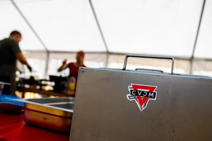 Eine Metallbox mit dem CVJM-Logo steht im Vordergrund vor einem unscharfen Hintergrund mit Personen unter einem weißen Zelt.