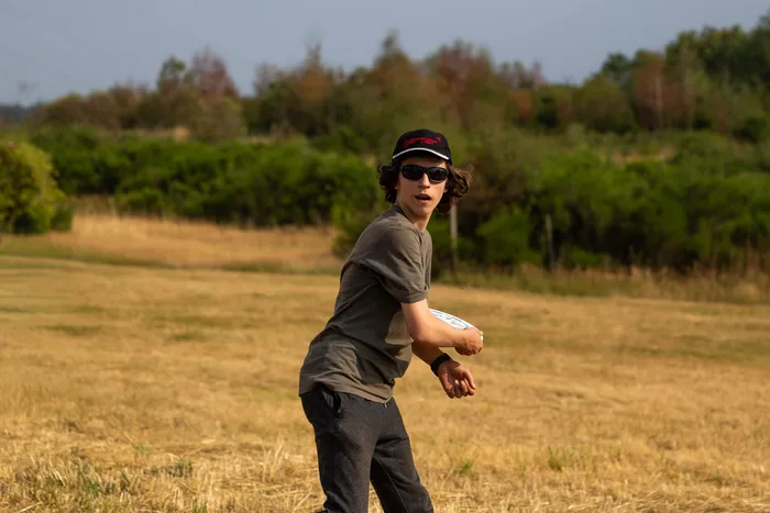 Ein junger Mann mit Sonnenbrille und schwarzer Kappe hält eine weiße Wurfscheibe auf einer trockenen Wiese.