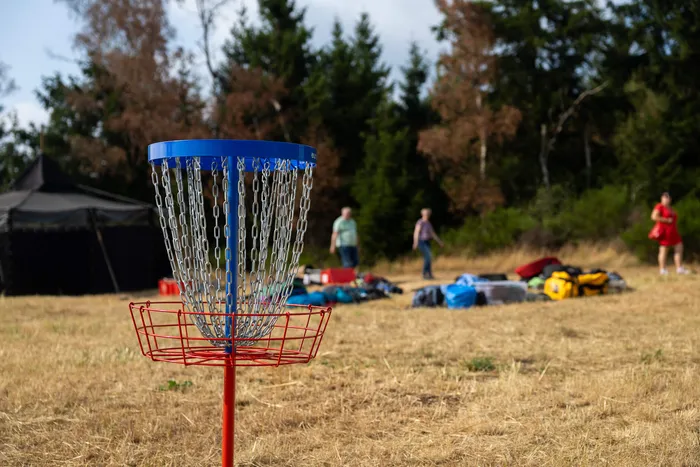 Ein Disc-Golf-Korb mit blauer Oberseite und rotem Korb steht auf einer trockenen Wiese vor einem Waldrand.