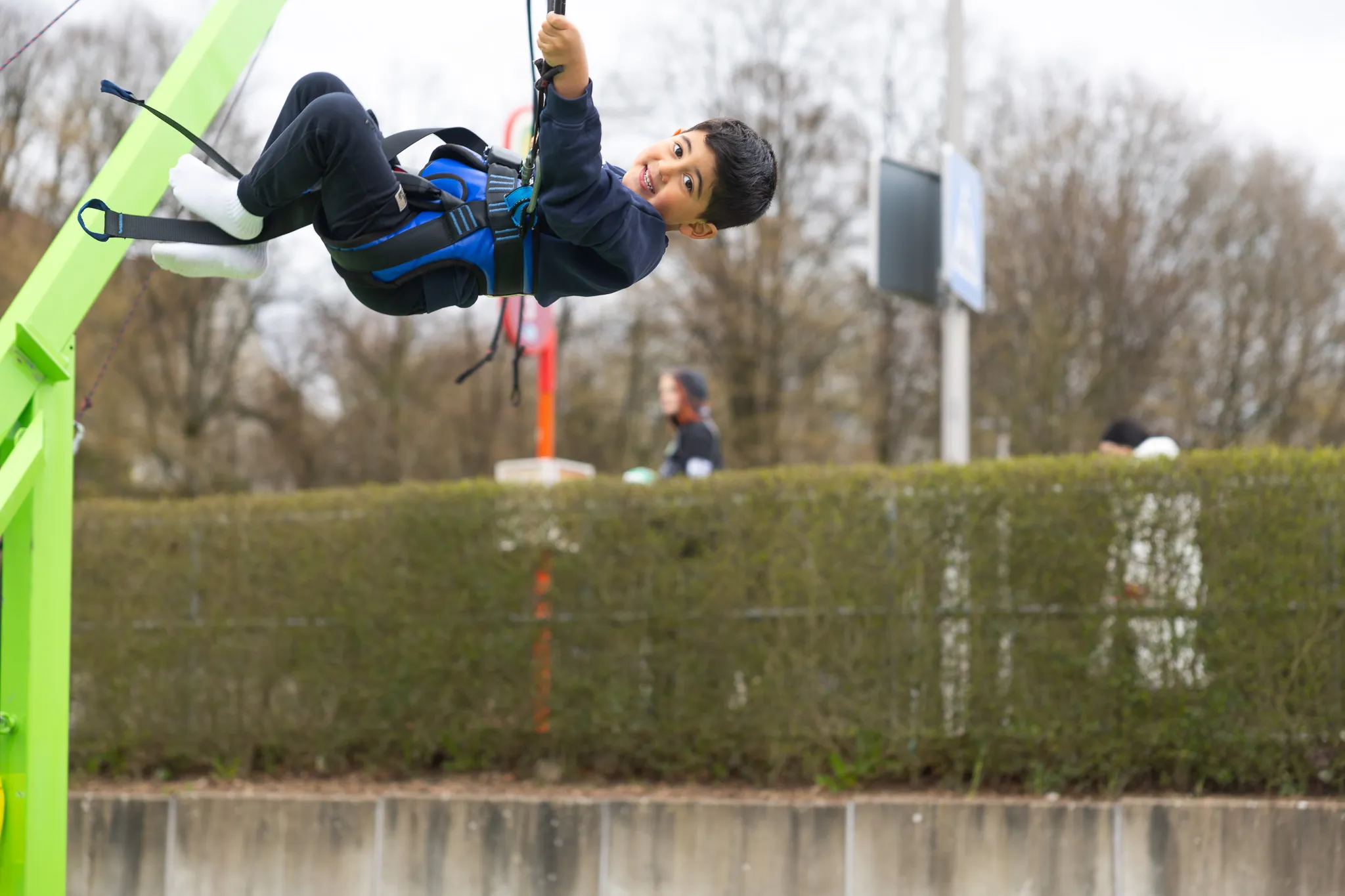Ein Junge mit dunklen Haaren und blauer Jacke schwingt an einer Seilrutsche, gesichert mit einem Gurt. Im Hintergrund ist eine grüne Hecke und eine verschwommene Person zu sehen.