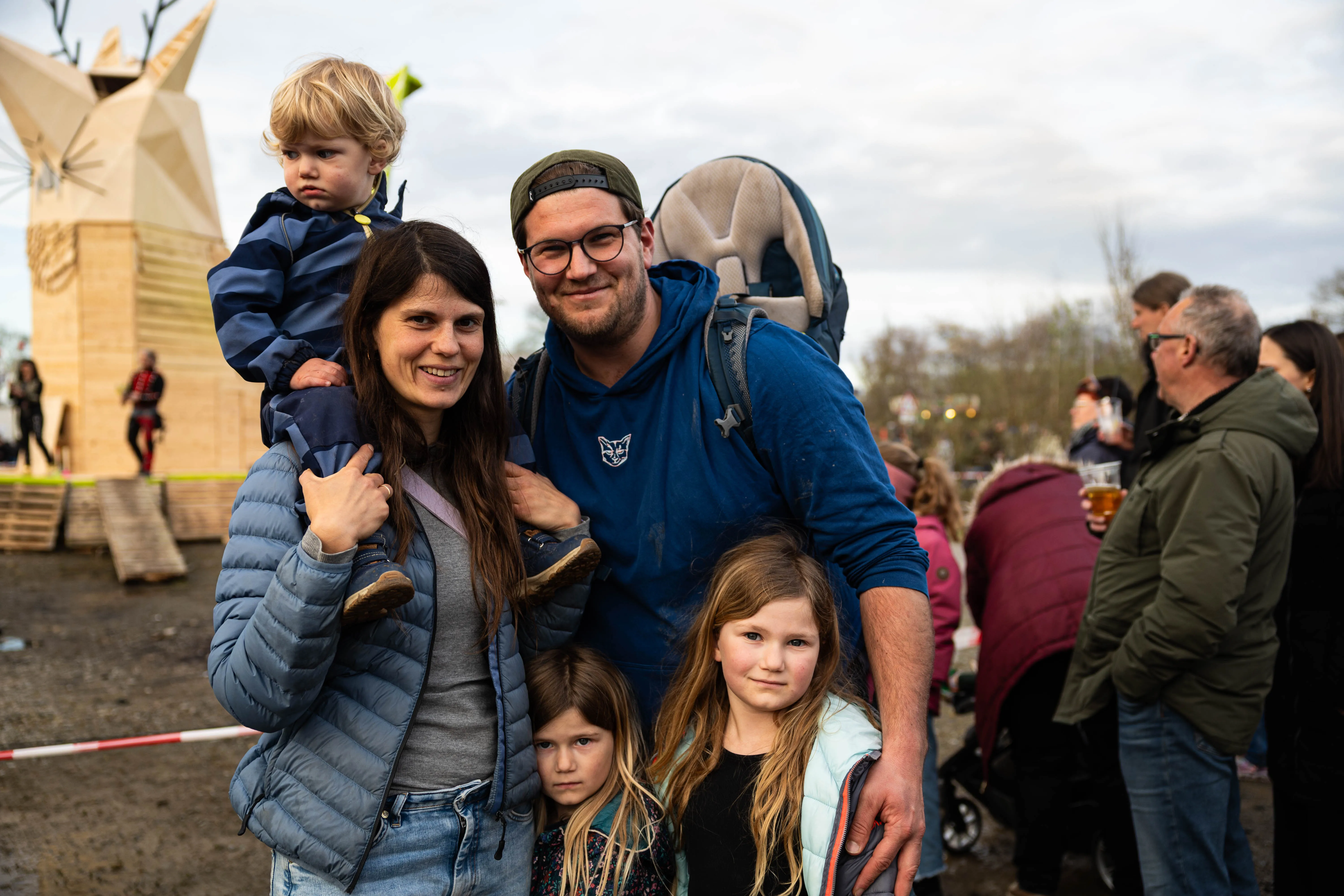 Familie mit drei Kindern lächelt in die Kamera, während sie auf einem Osterfeuer-Gelände steht. Im Hintergrund sind weitere Besucher und eine skulpturale Holzkonstruktion zu sehen.