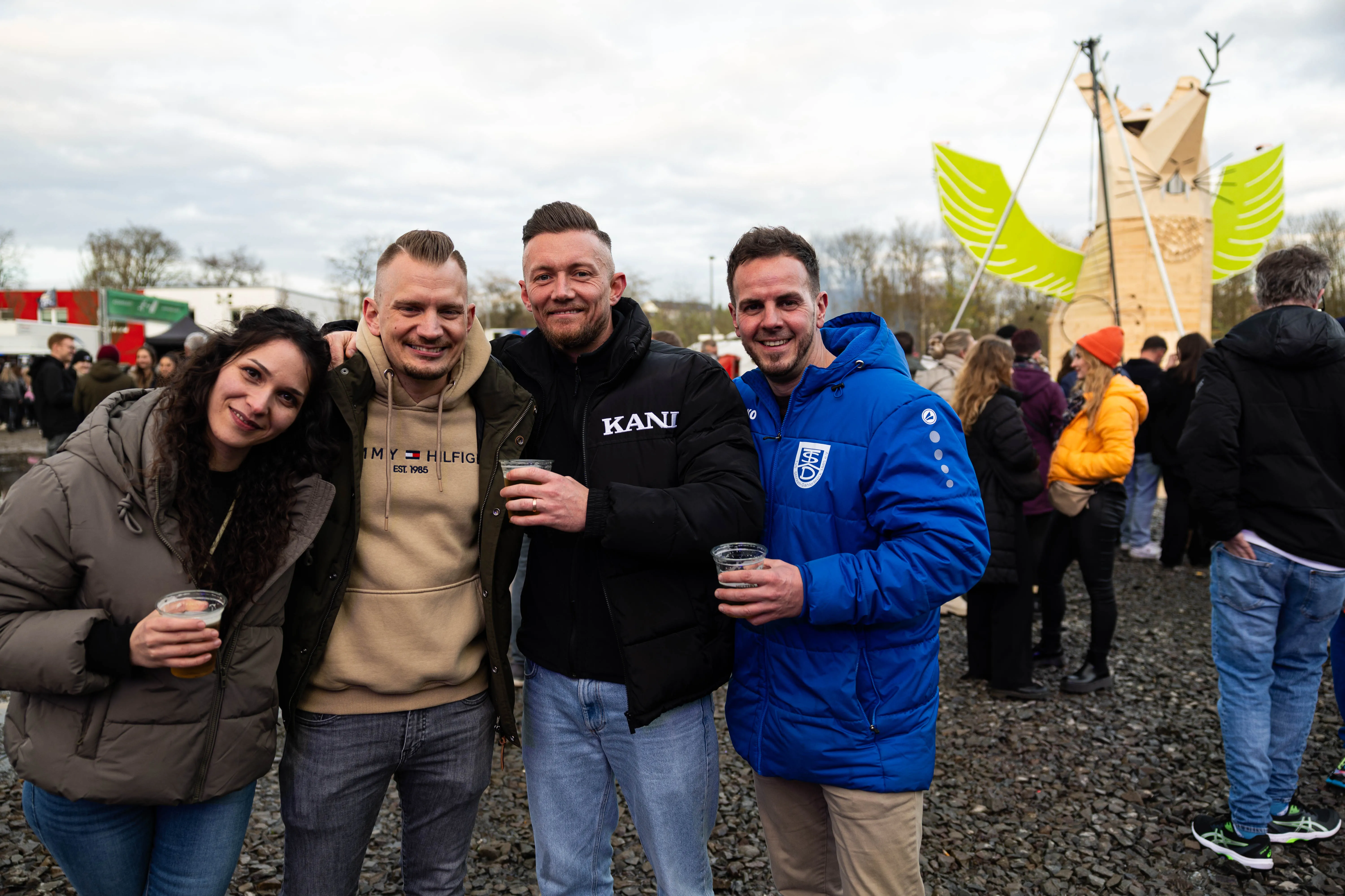 Vier junge Erwachsene stehen lächelnd vor der Kamera und halten jeweils ein Glas in der Hand. Im Hintergrund sind weitere Menschen und eine große Holzkonstruktion zu sehen.