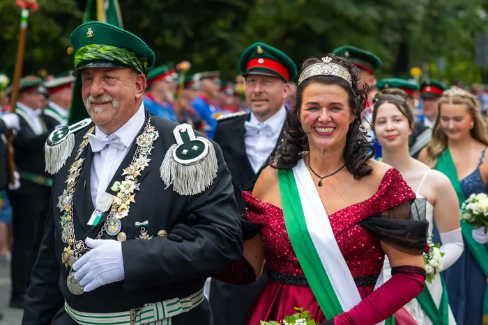 Stefan Schneider in schwarzer Schützenuniform mit grüner Mütze und Orden geht arm in arm mit einer Frau in rotem Kleid und weiß-grüner Schärpe.