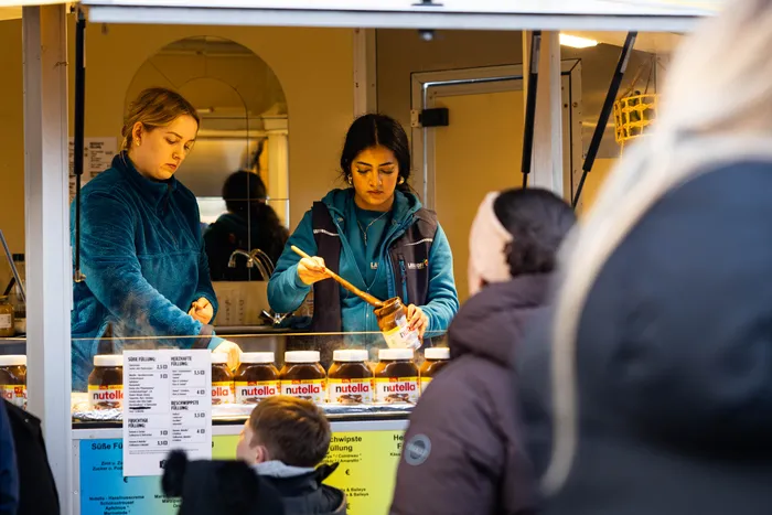 Zwei Frauen bereiten in einem Imbisswagen Crêpes zu, während im Hintergrund Kunden stehen.