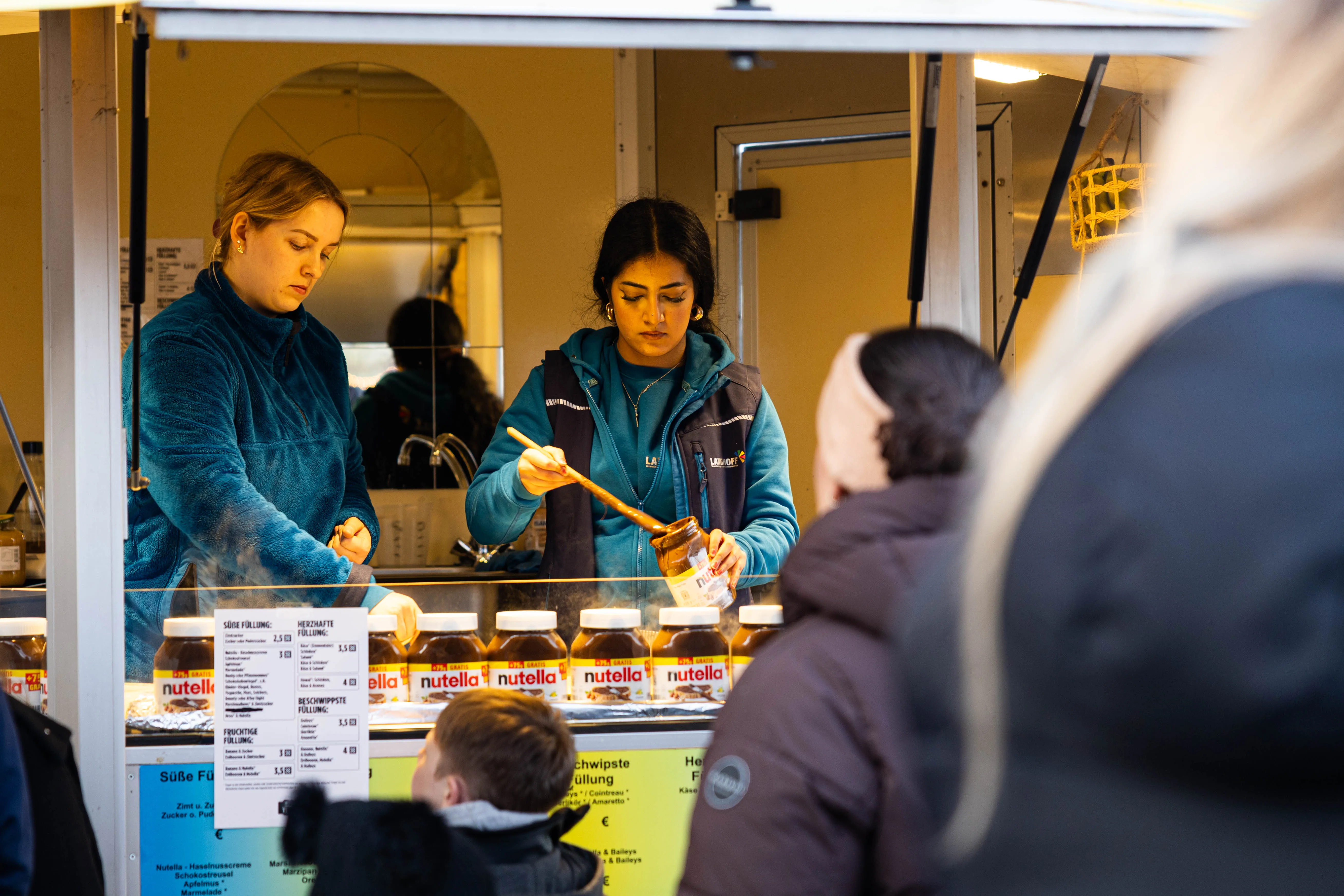Zwei Frauen bereiten in einem Imbisswagen Crêpes zu, während im Hintergrund Kunden stehen.