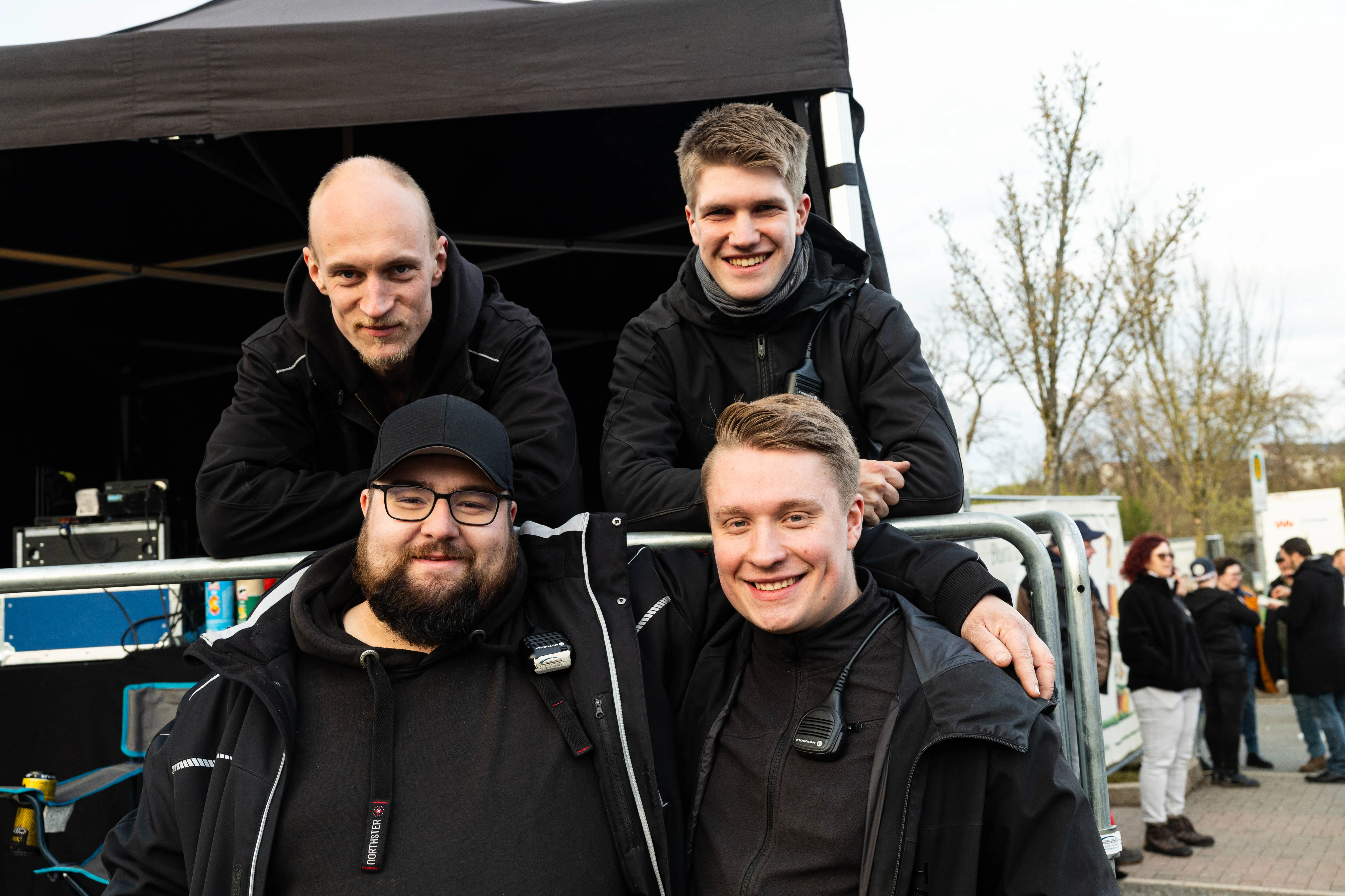 Vier lächelnde Männer posieren eng zusammen vor einer Bühne und Zuschauern bei einem Osterfeuer. Zwei tragen Kapuzenjacken, einer eine Baseballkappe.