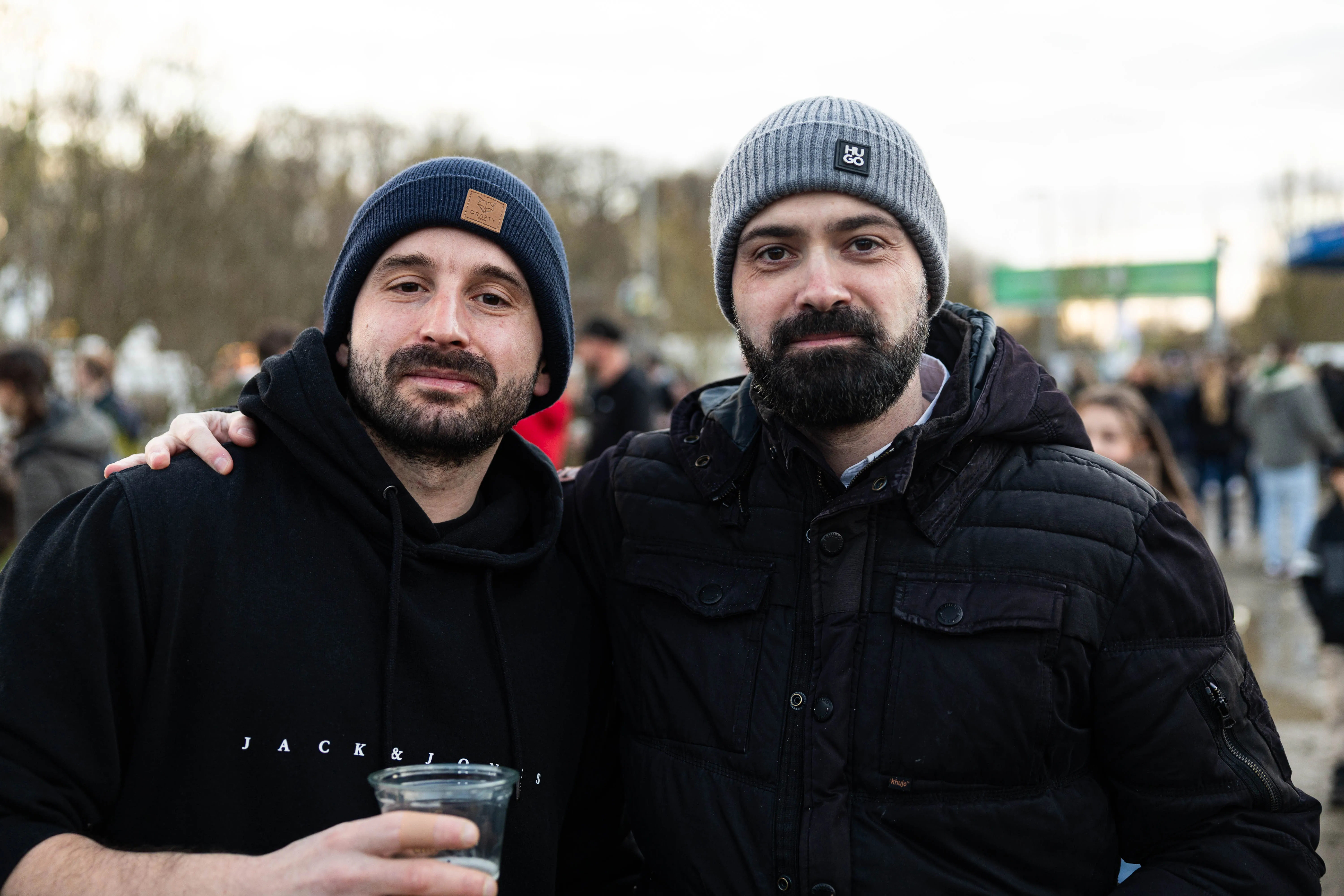 Zwei Männer mit Mützen und Bart stehen eng zusammen vor einer Menschenmenge. Einer hält ein Glas in der Hand.