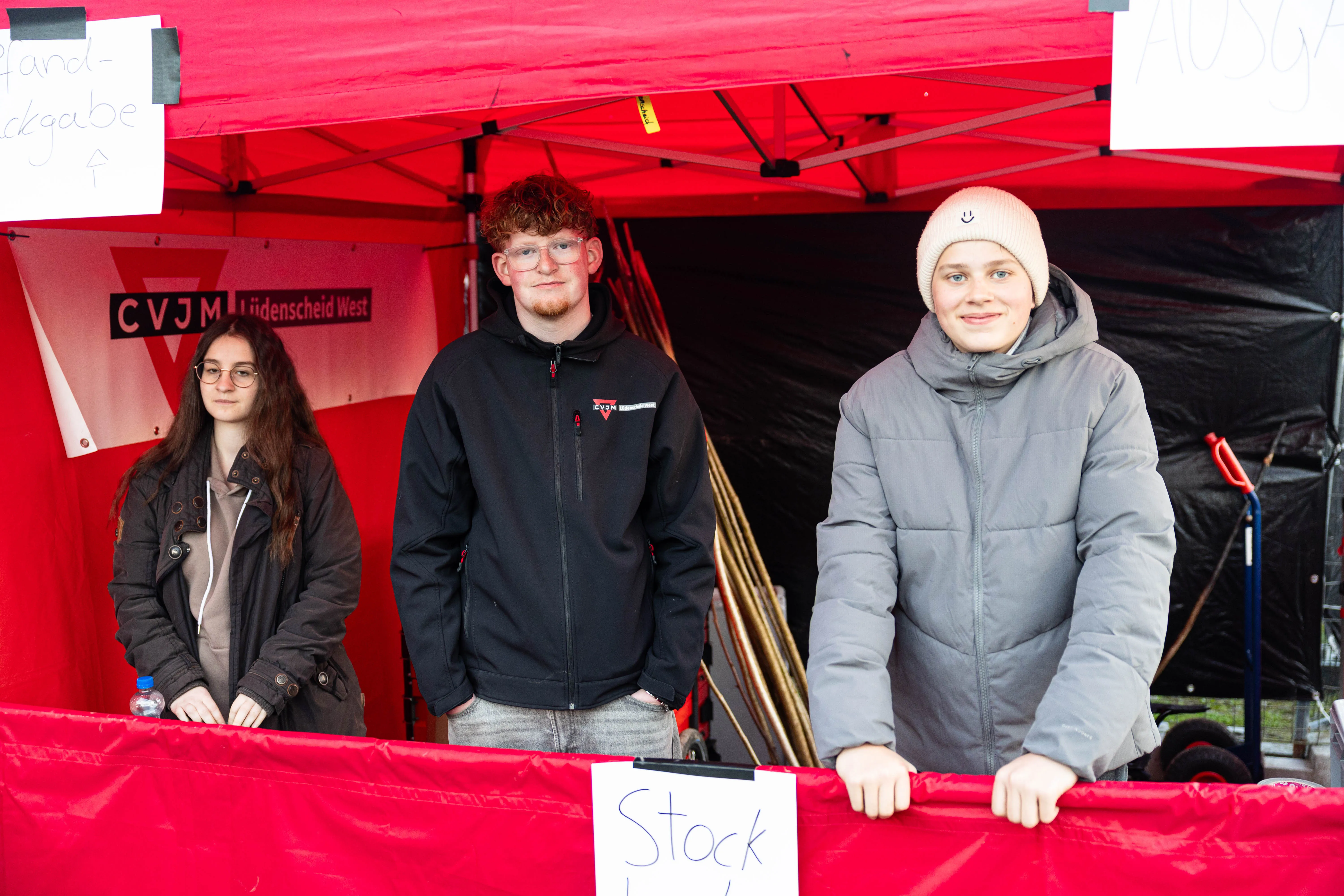 Drei junge Erwachsene stehen hinter einem roten Tisch mit der Aufschrift „Stock“ vor einem roten Zelt mit dem Logo CVJM Lüdenscheid West.