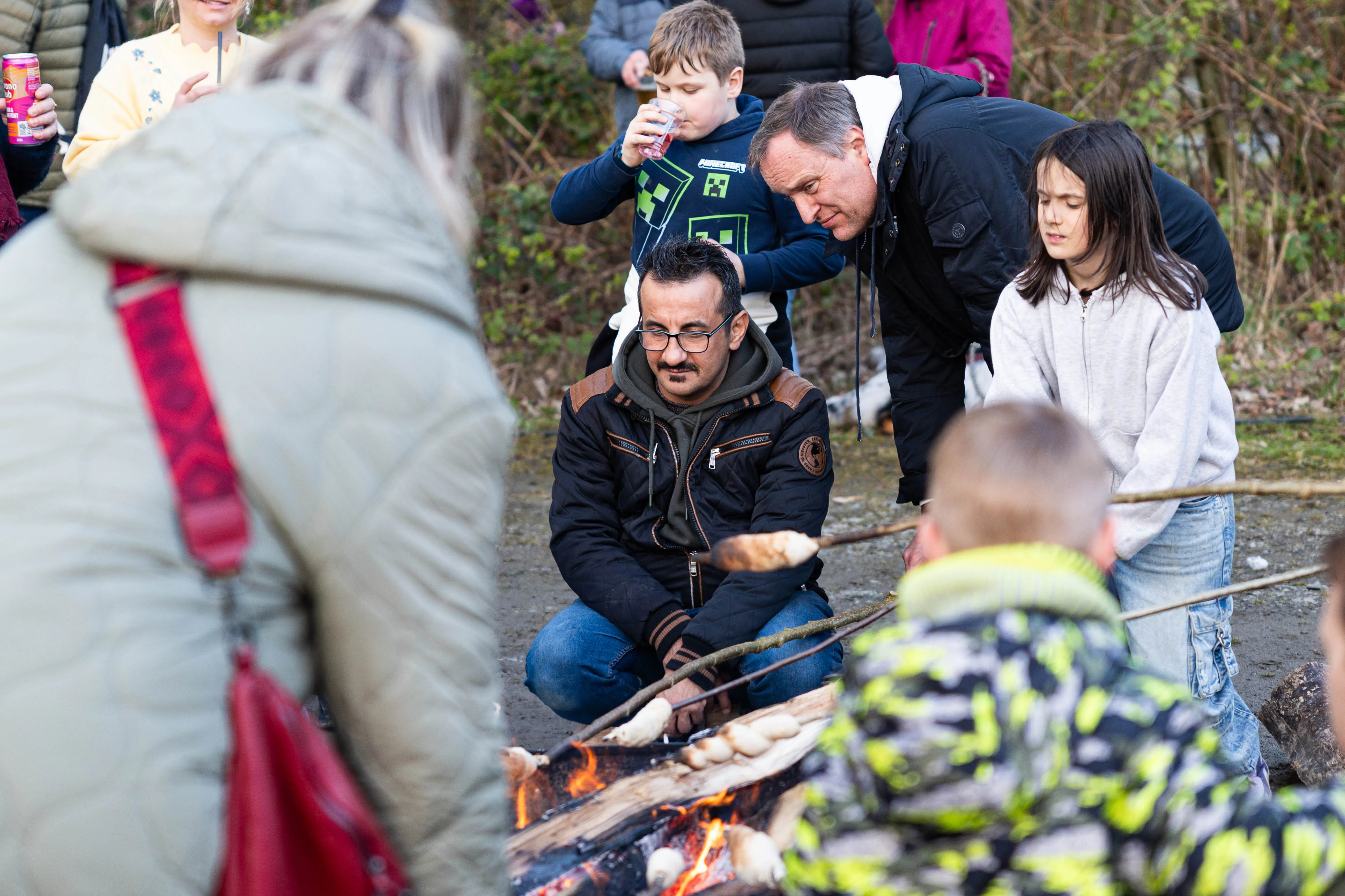 Menschen stehen und hocken um ein Lagerfeuer, auf dem Marshmallows geröstet werden. Im Hintergrund sind weitere Personen und Büsche zu sehen.