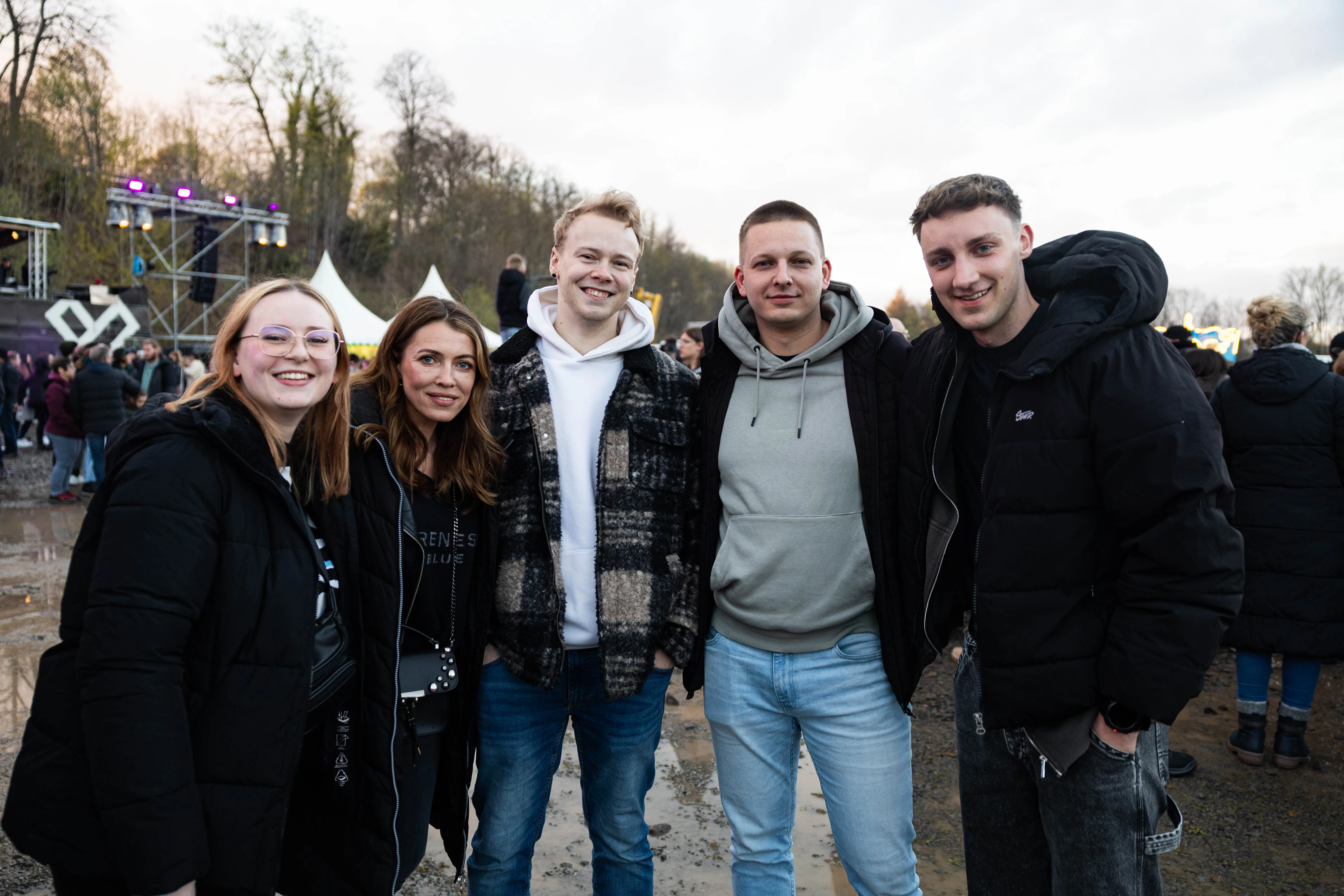 Fünf junge Erwachsene stehen lächelnd vor einer Bühne und Zuschauermassen auf dem Gelände des Osterfeuers „Burn the Fox“ in Lüdenscheid.