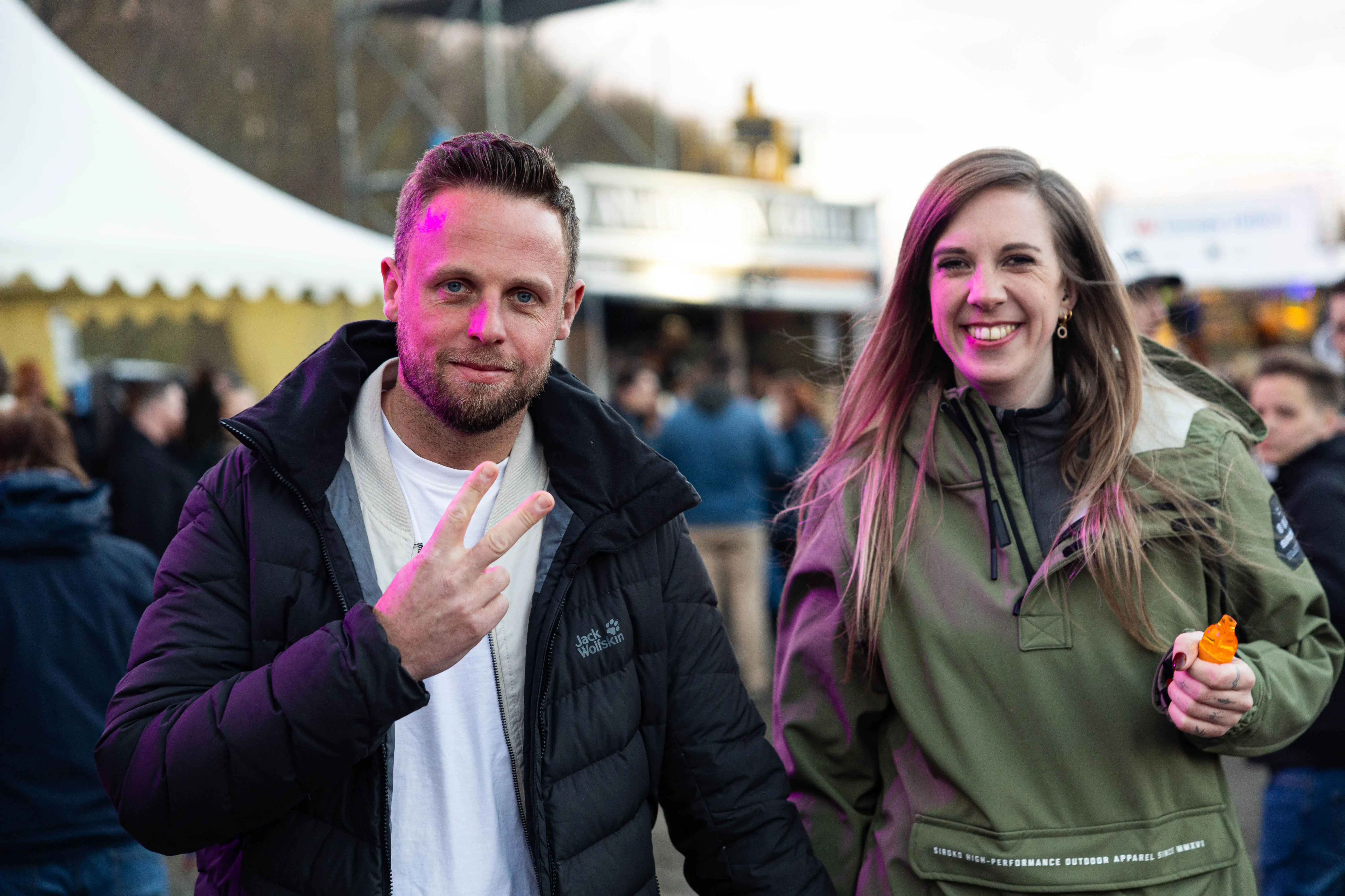 Zwei lächelnde Menschen vor einer Menschenmenge auf einem Festivalgelände. Der Mann macht ein Friedenszeichen, beide tragen Winterkleidung.