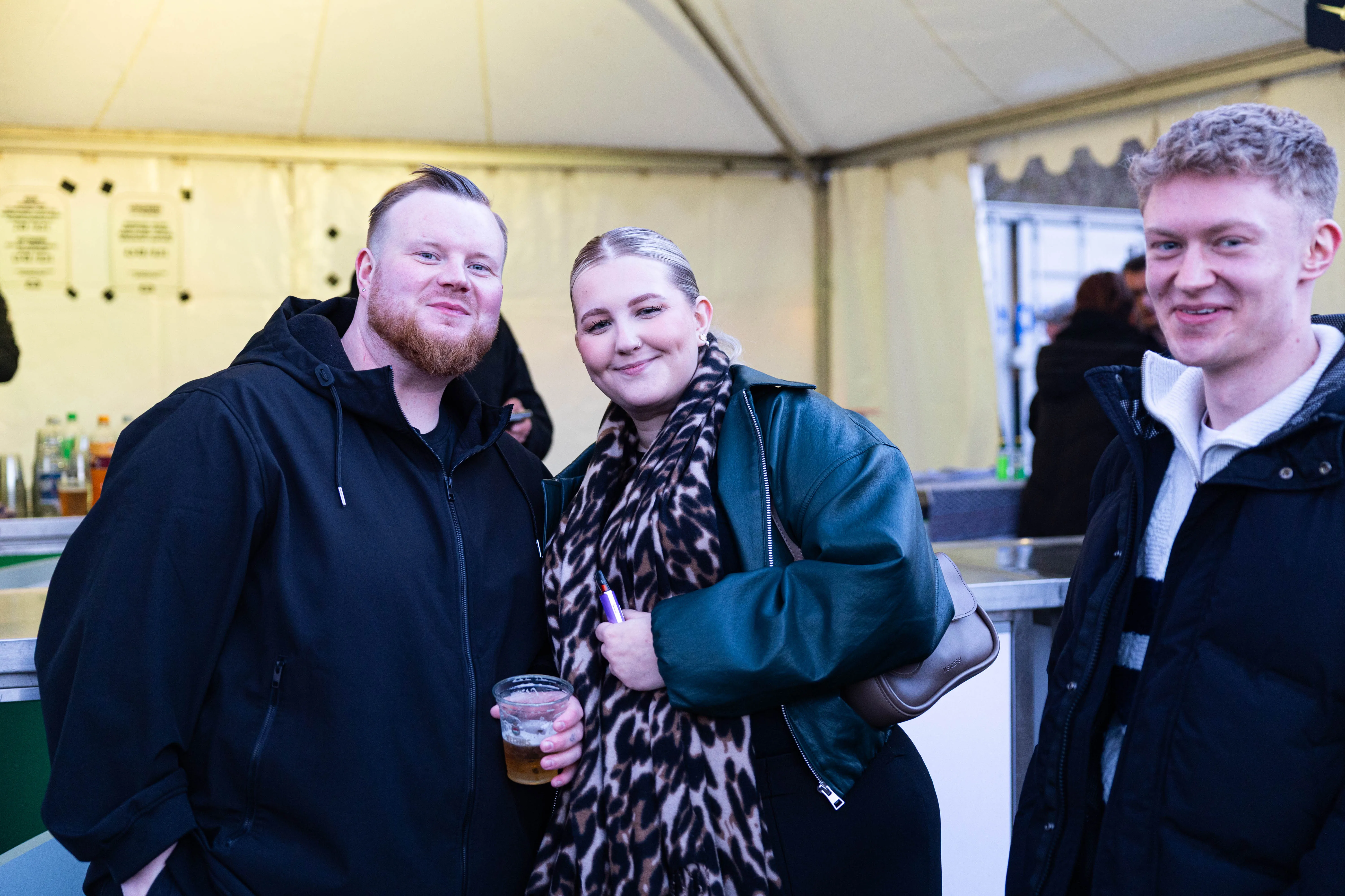 Drei junge Erwachsene lächeln in die Kamera, vor ihnen eine Bar unter einem Zelt. Im Hintergrund sind weitere Personen zu sehen.